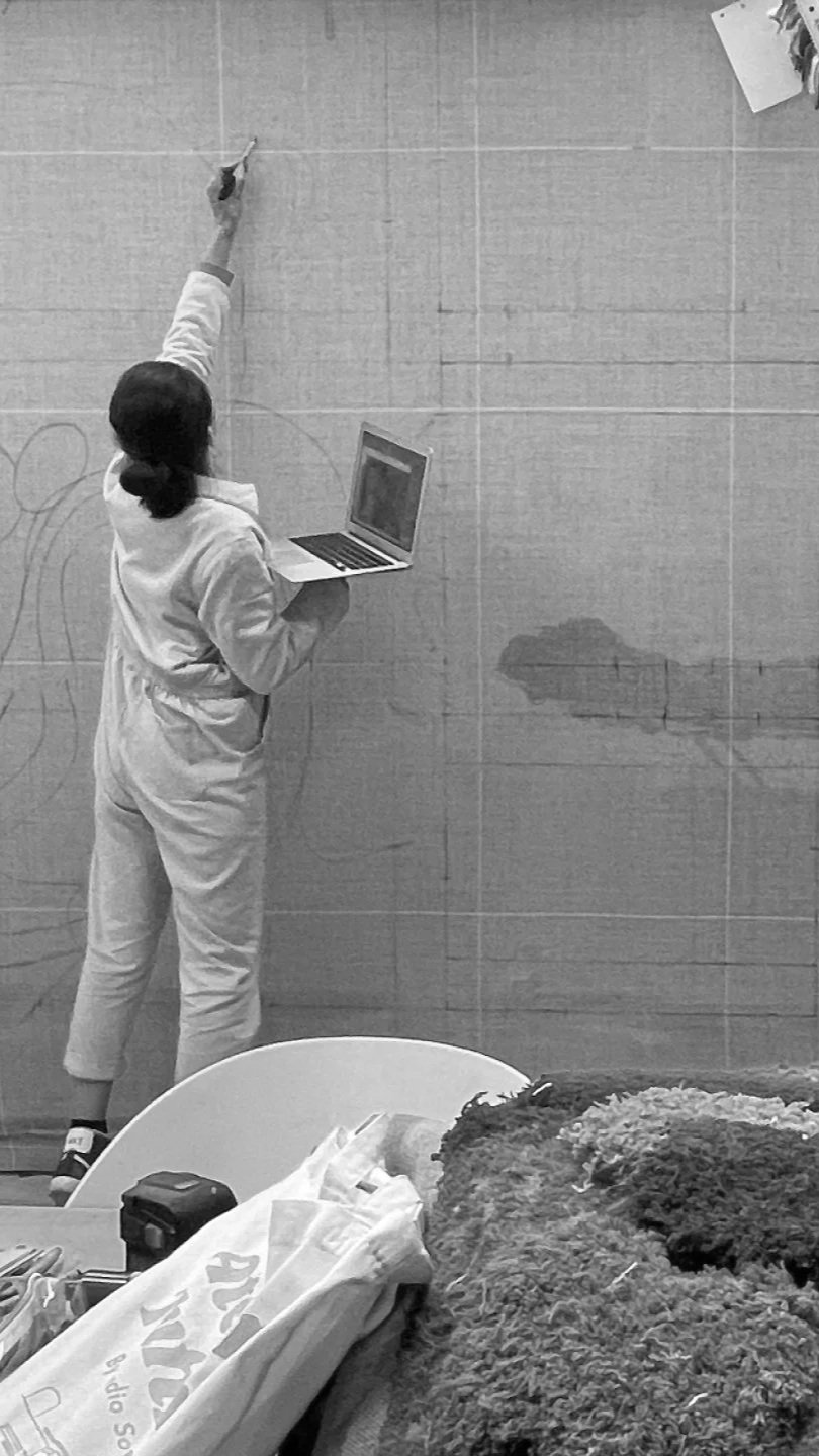 A person in a white scrub suit drawing on a wall with a marker, while holding a laptop. The wall has faint outlines of shapes and patterns. There are clothes and objects on a table in the foreground.