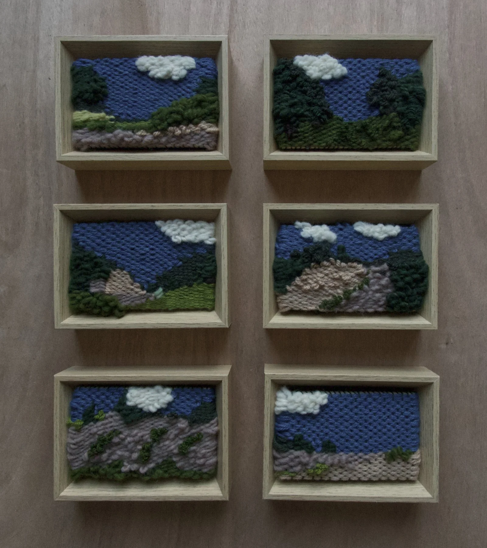 Six small framed textile landscape art pieces arranged in two columns and three rows on a wooden surface, with mountains, trees, sky, and clouds depicted in textured yarn.