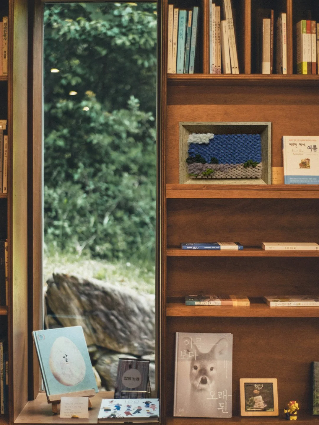 Bookshelves with books and a framed embroidered landscape art in a bookstore or library, with a large window showing trees and outdoor greenery.