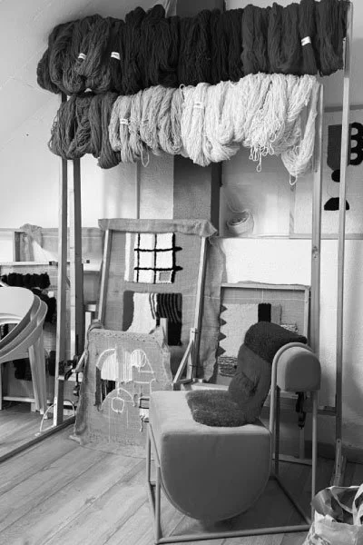 Display of large skeins of yarn in black, white, and gray on a metal rack, with a chair and fabric wall hangings nearby.