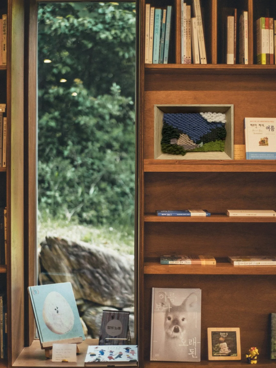 Books on wooden shelves beside a large window showing green trees outside. A framed textile art piece depicts mountains, sky, and trees. Additional artwork and books are displayed on a wooden table below the shelves.
