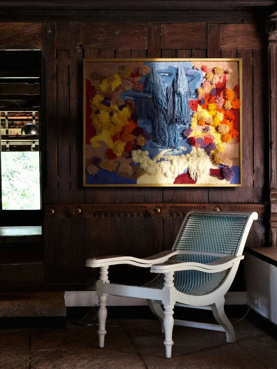 A white wooden chair with a woven cane seat and backrest, placed in front of a dark wooden wall with a colorful, textured embroidery artwork hanging on it.