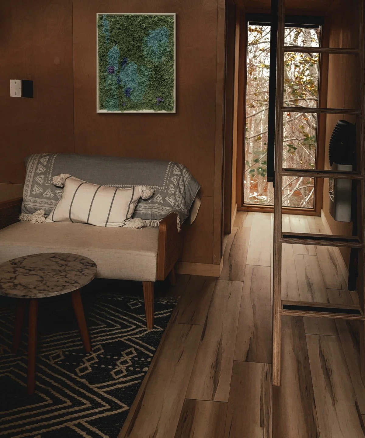 A cozy living room corner with a beige sofa adorned with striped pillows, a round marble-top coffee table, a wooden wall with a framed green moss art piece, and a sliding glass door revealing autumnal trees outside.