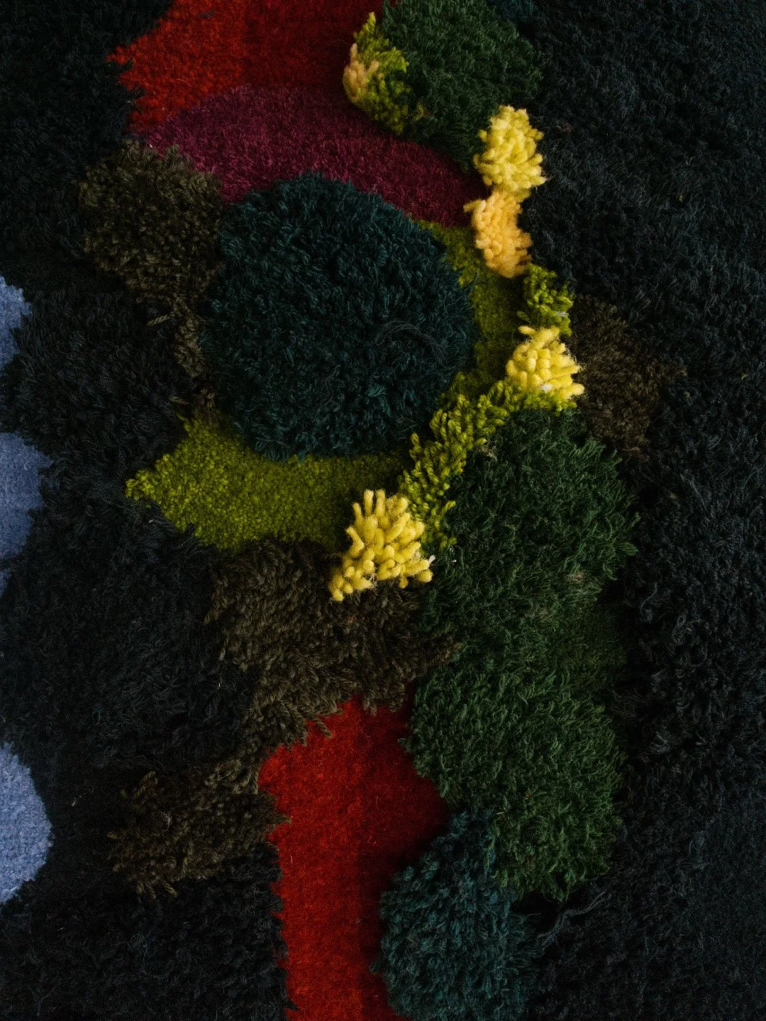 Close-up of a textured, multicolored, knitted or crocheted design with various sections of different colors including black, green, yellow, red, and purple, featuring bumpy, pom-pom and looped textures.