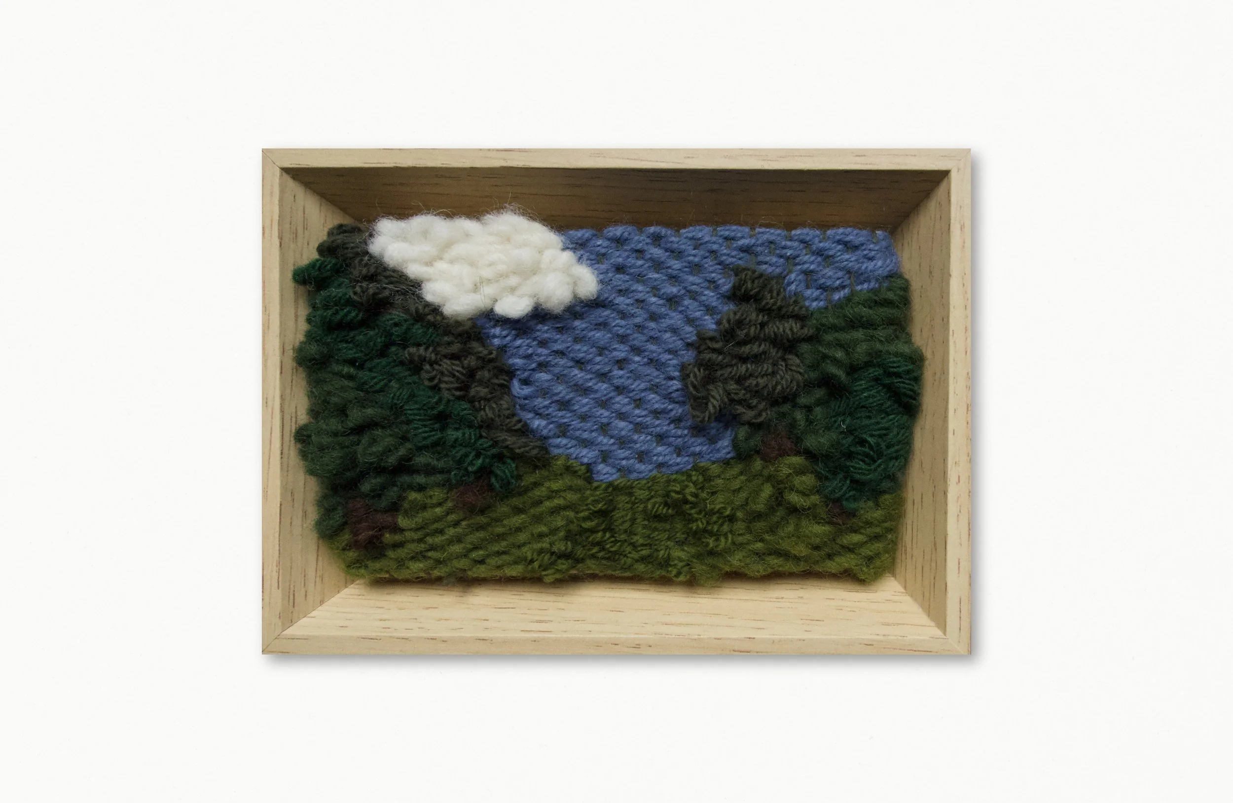Textured embroidery piece with a blue sky, white cloud, green trees, and dark brown mountains in a wooden frame.