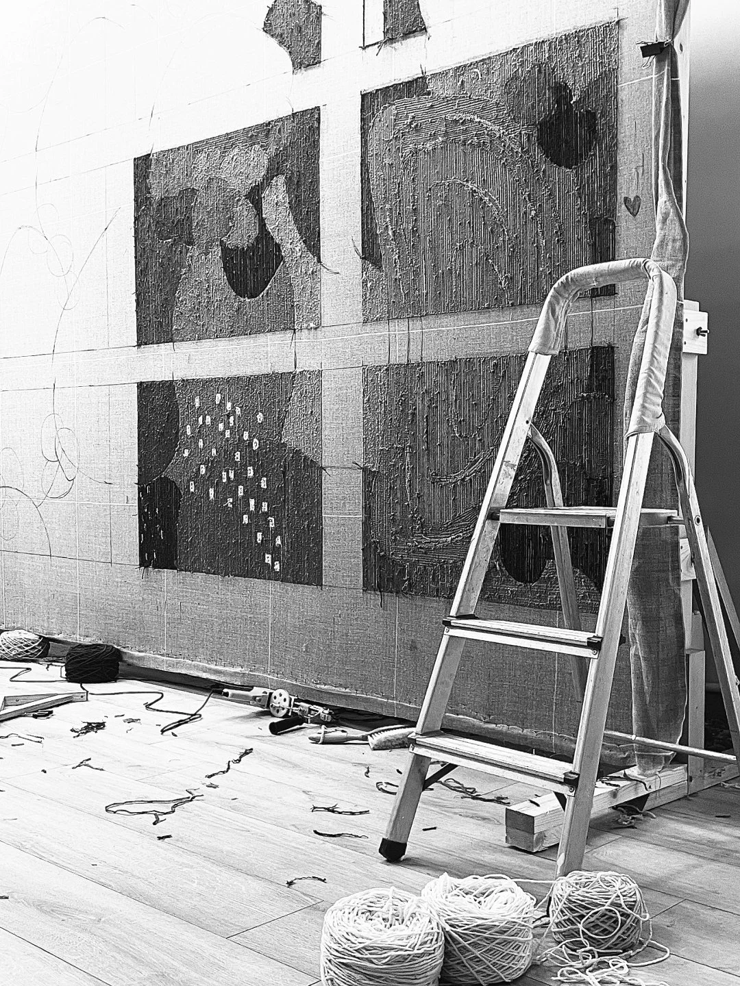 A wall with four square panels of artwork in progress, a step ladder, and art supplies including yarn and tools on the floor.