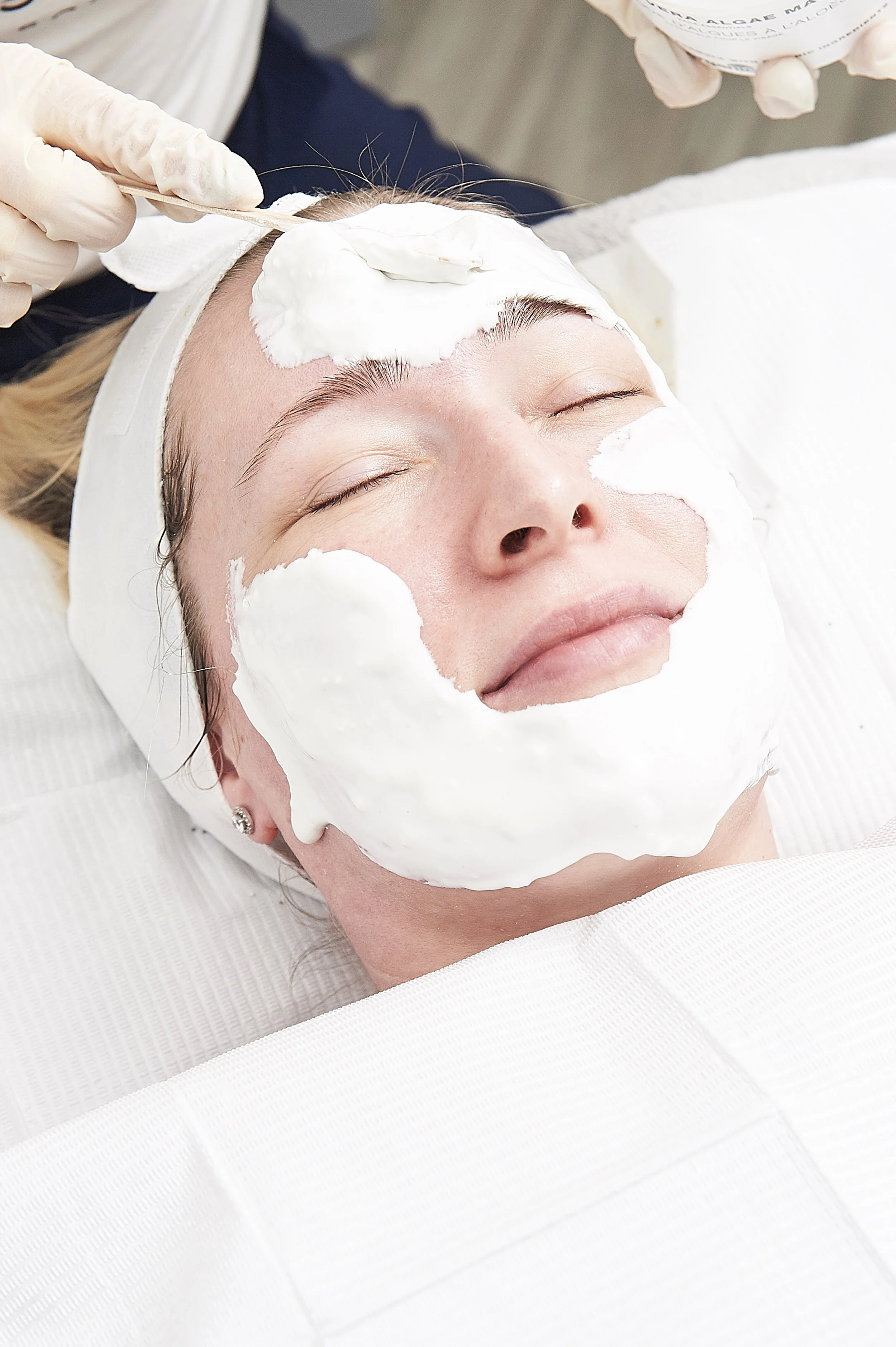 A woman lying down with her eyes closed while receiving a facial treatment with white foam or mask applied to her face.