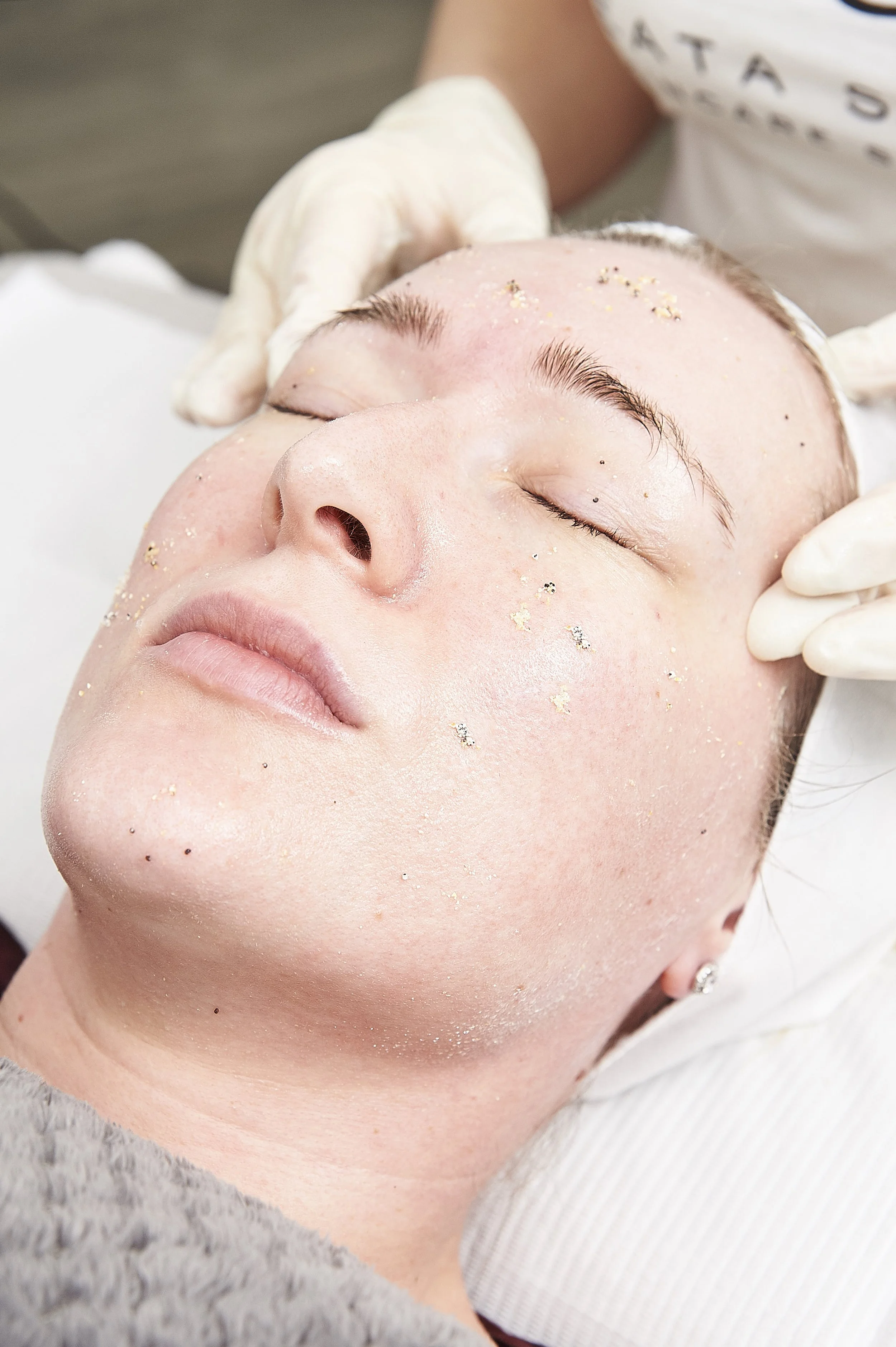 A woman receiving a facial treatment with small exfoliating beads or particles on her face, lying with her eyes closed while a skincare professional in gloves works on her.