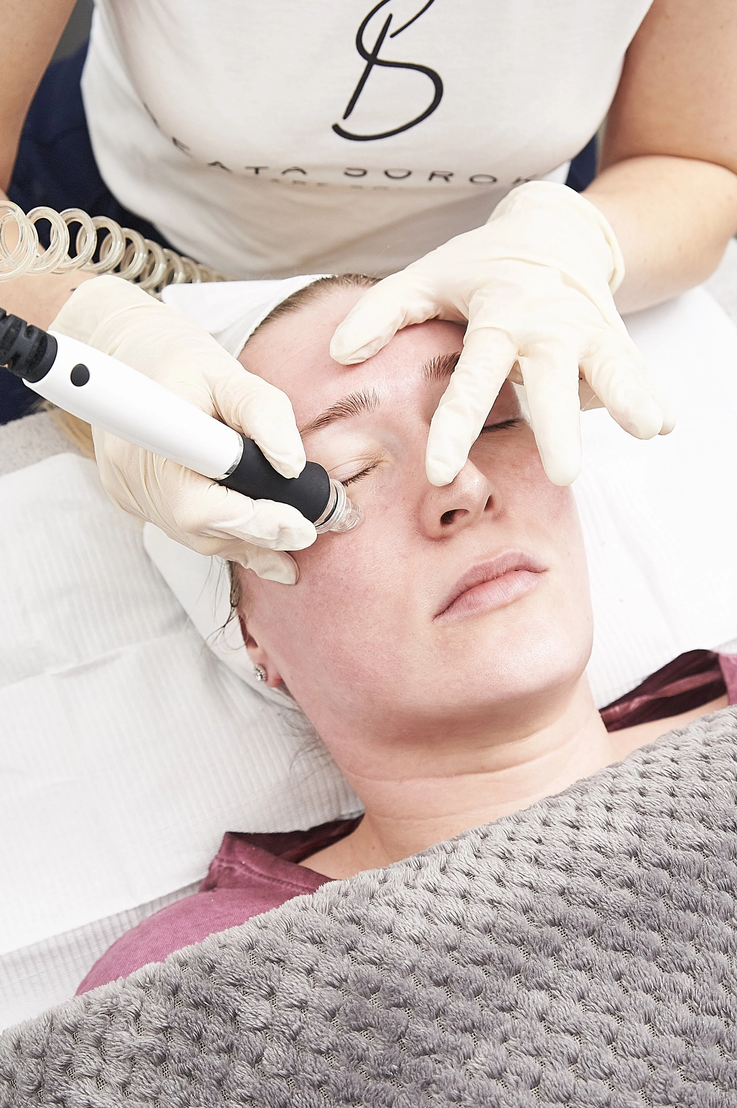 A woman receiving a facial treatment with a handheld device, lying on a treatment bed with a textured blanket, while a technician in gloves applies the procedure.