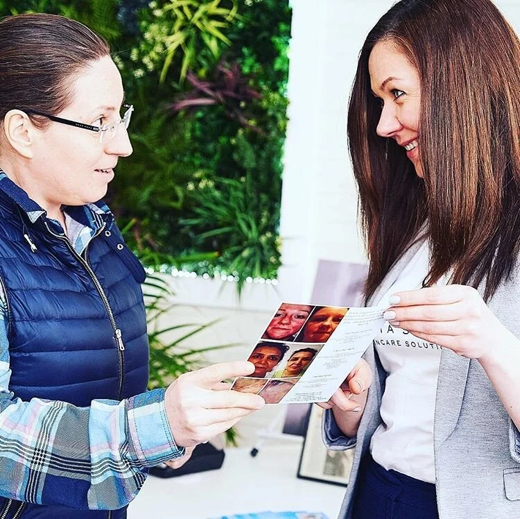Two women are standing indoors, one is showing the other a brochure or flyer with pictures of faces and skincare solutions. They are smiling and appear to be engaged in a friendly conversation.