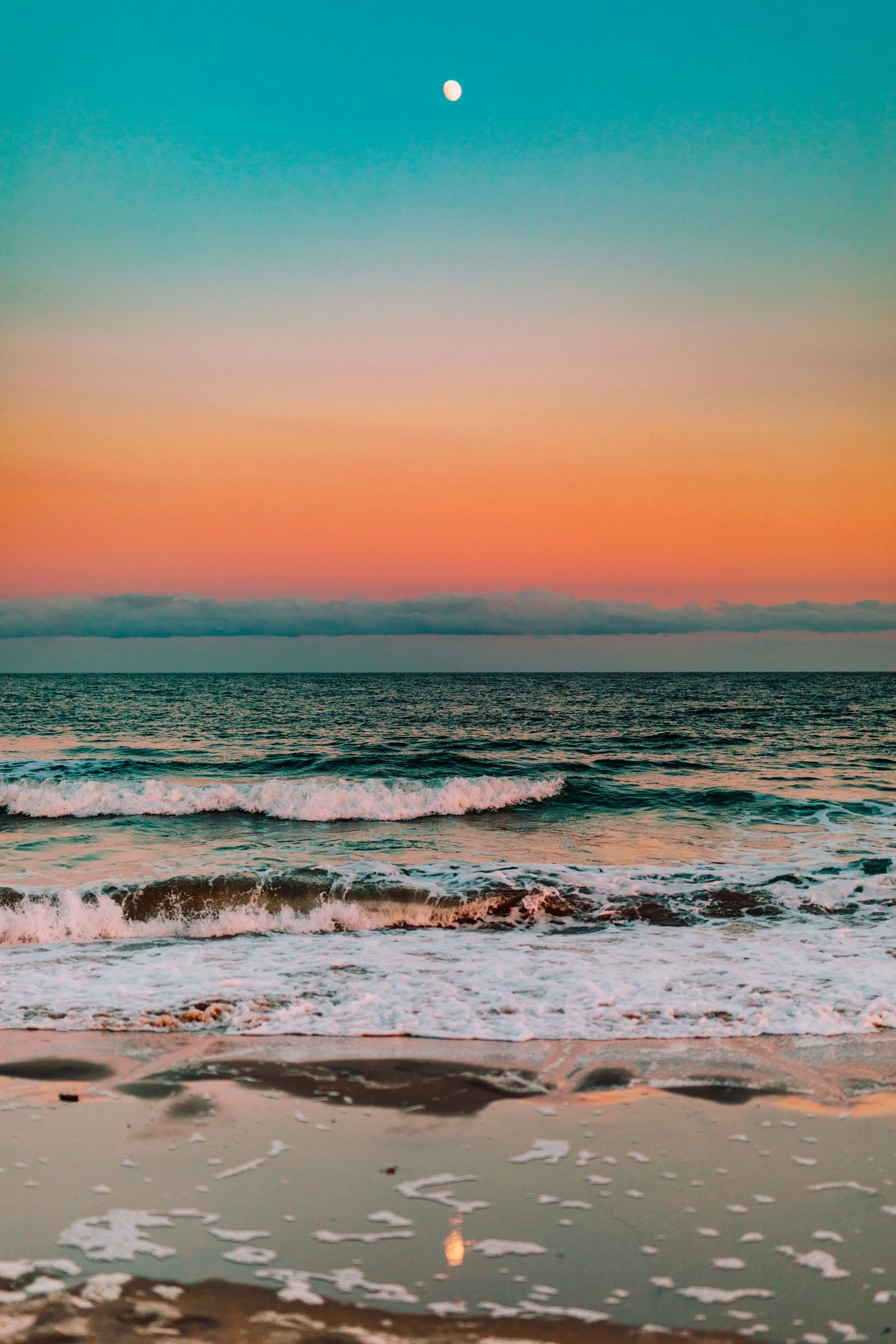 A beach at sunset with waves crashing onto the sand and the moon visible in the sky.