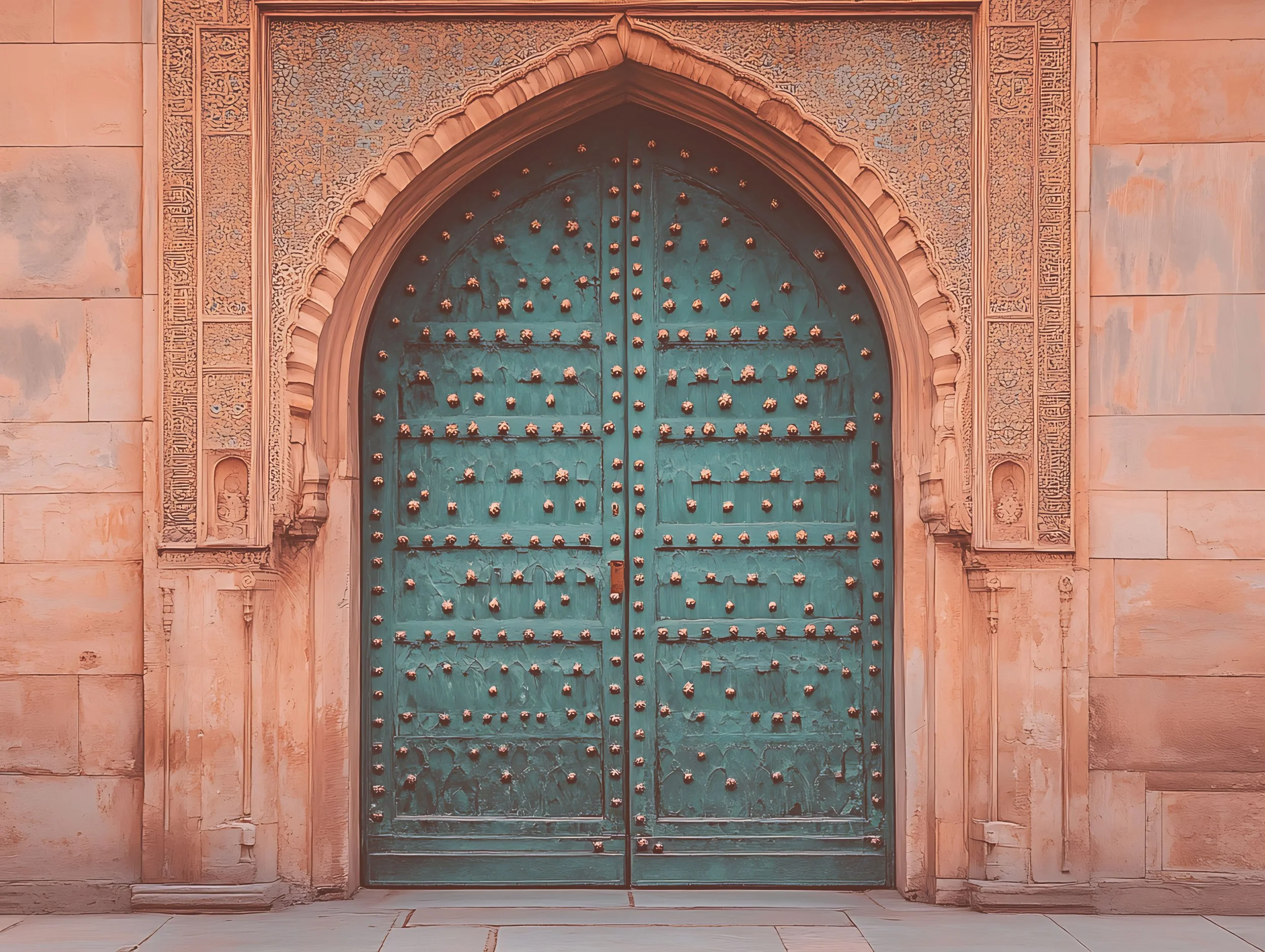 A large ornate teal-colored metal door with decorative rivets, set within a stone archway with intricate carvings, on a peach-colored stone wall.