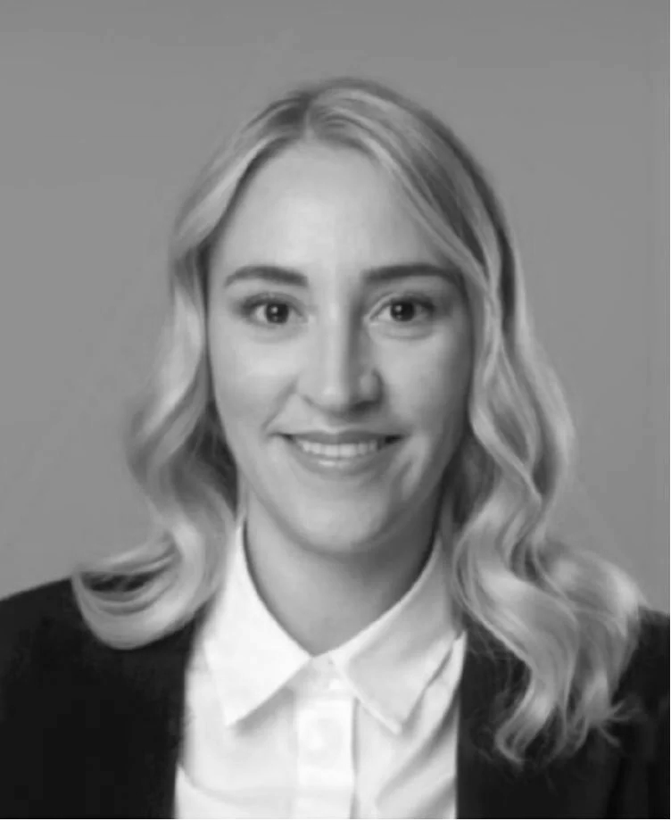 Schwarz-Weiß-Foto einer lächelnden Frau mit gewelltem, schulterlangem Haar, weißem Hemd und dunklem Blazer vor hellem Hintergrund.