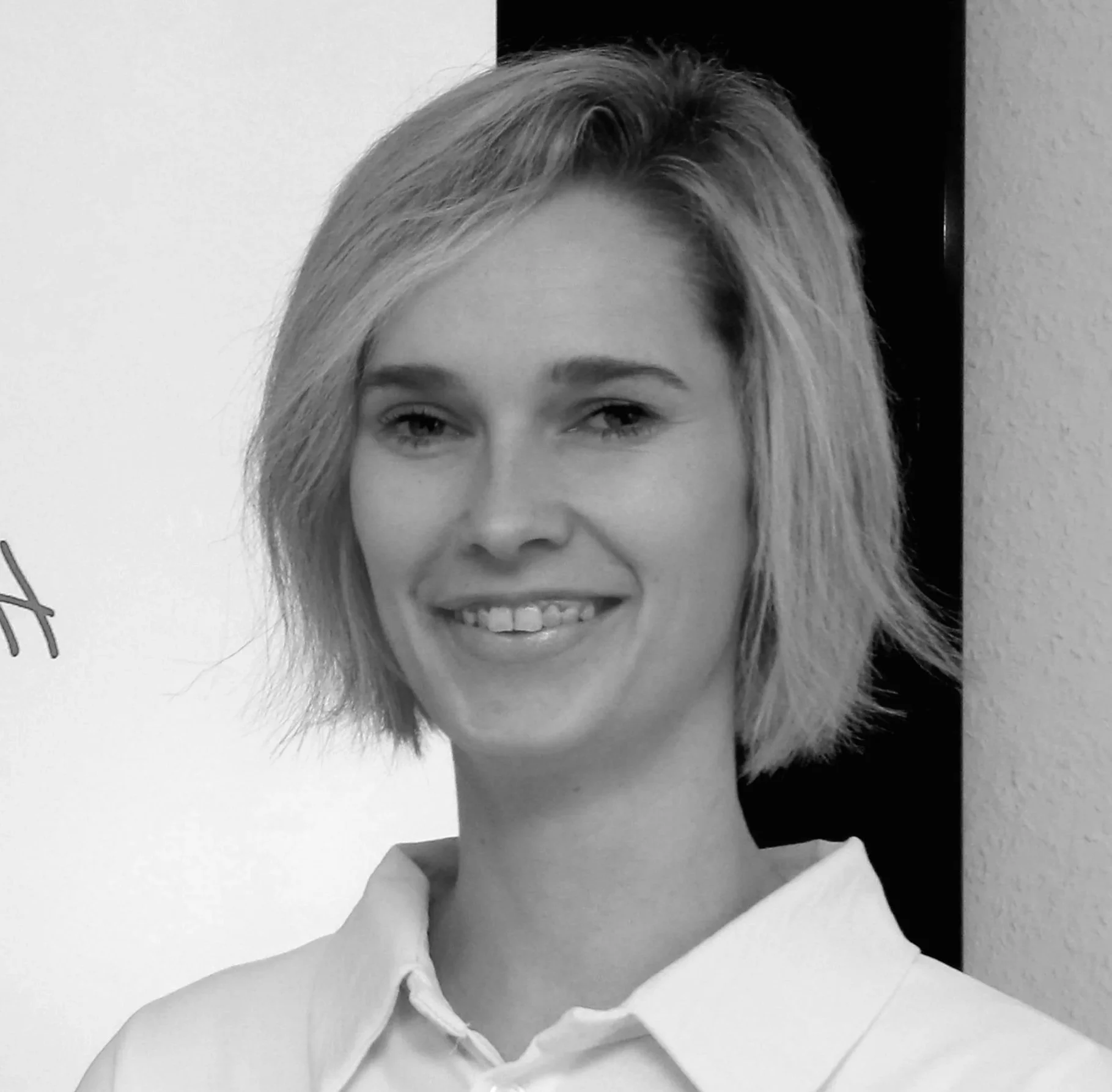 Schwarze und weiße Portraitaufnahme einer jungen Frau mit kurzem, welligem Haar, die lächelt, vor einer weißen Wand und dunklem Hintergründe.