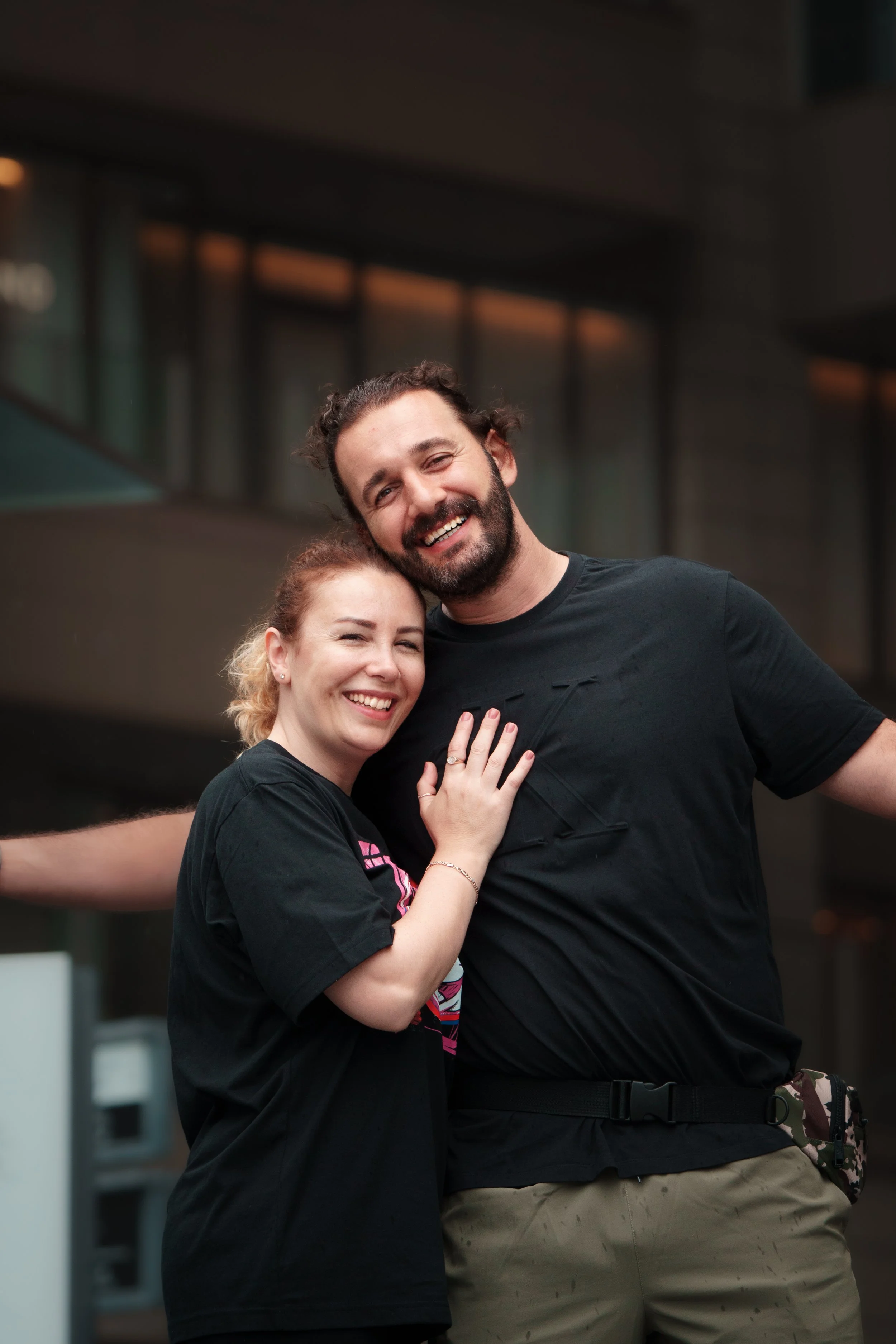 A woman and a man smiling, hugging each other outdoors, with a modern building in the background.