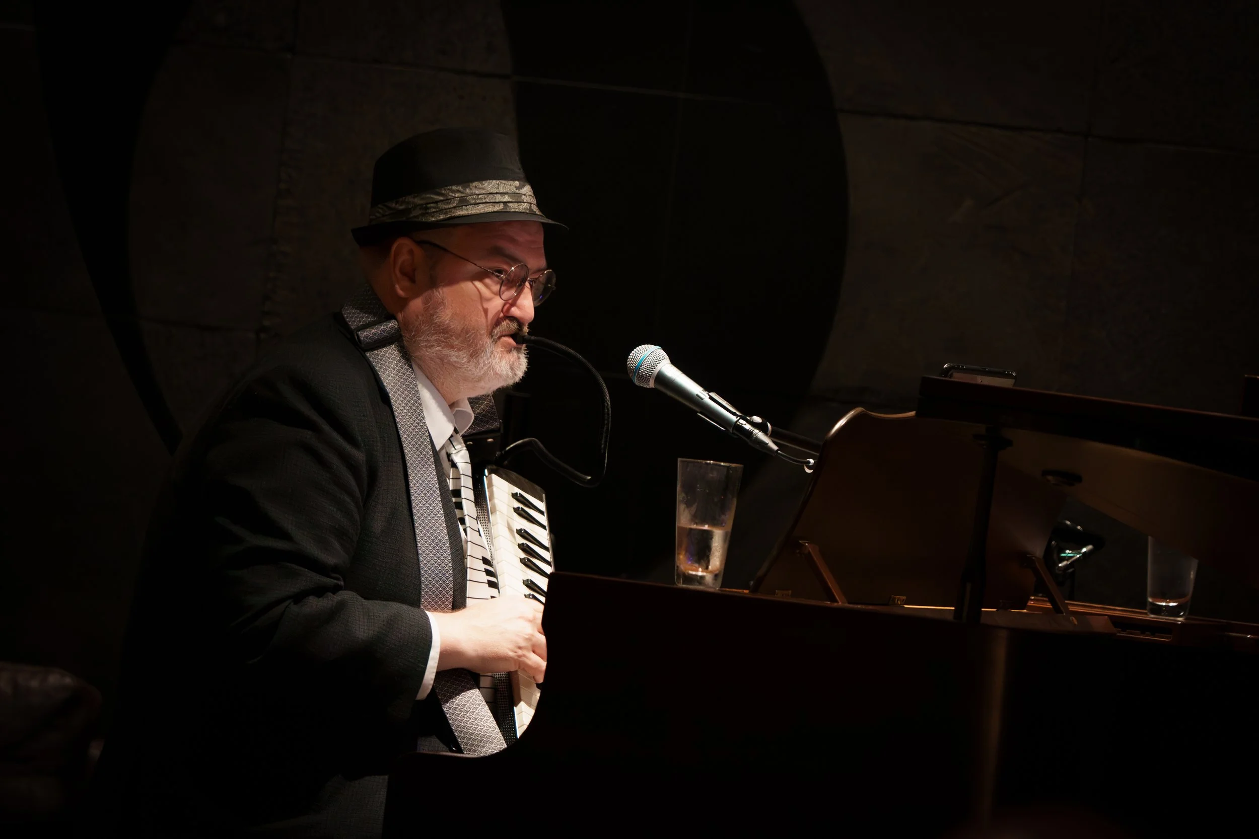 A man wearing glasses and a dark hat plays a grand piano at a microphone, with a glass of water nearby, in a dimly lit setting.