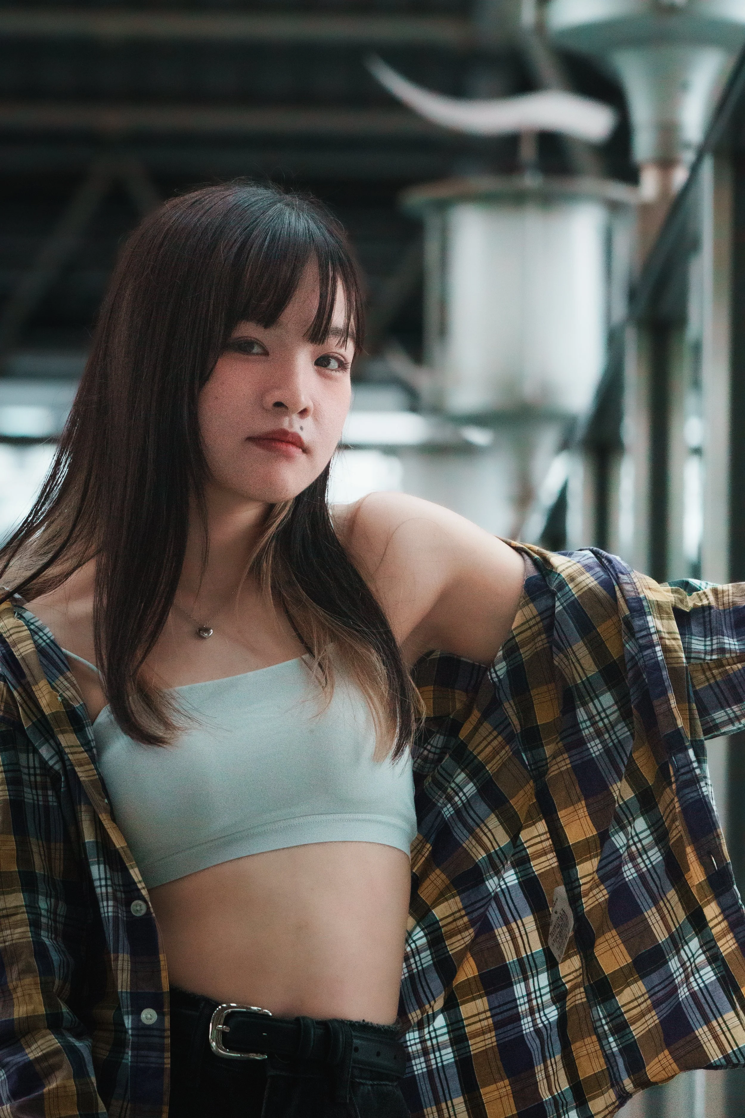 A young woman with dark hair and bangs looking at the camera, wearing a white crop top and a plaid shirt open over it, standing indoors with industrial pipes and windows in the background.