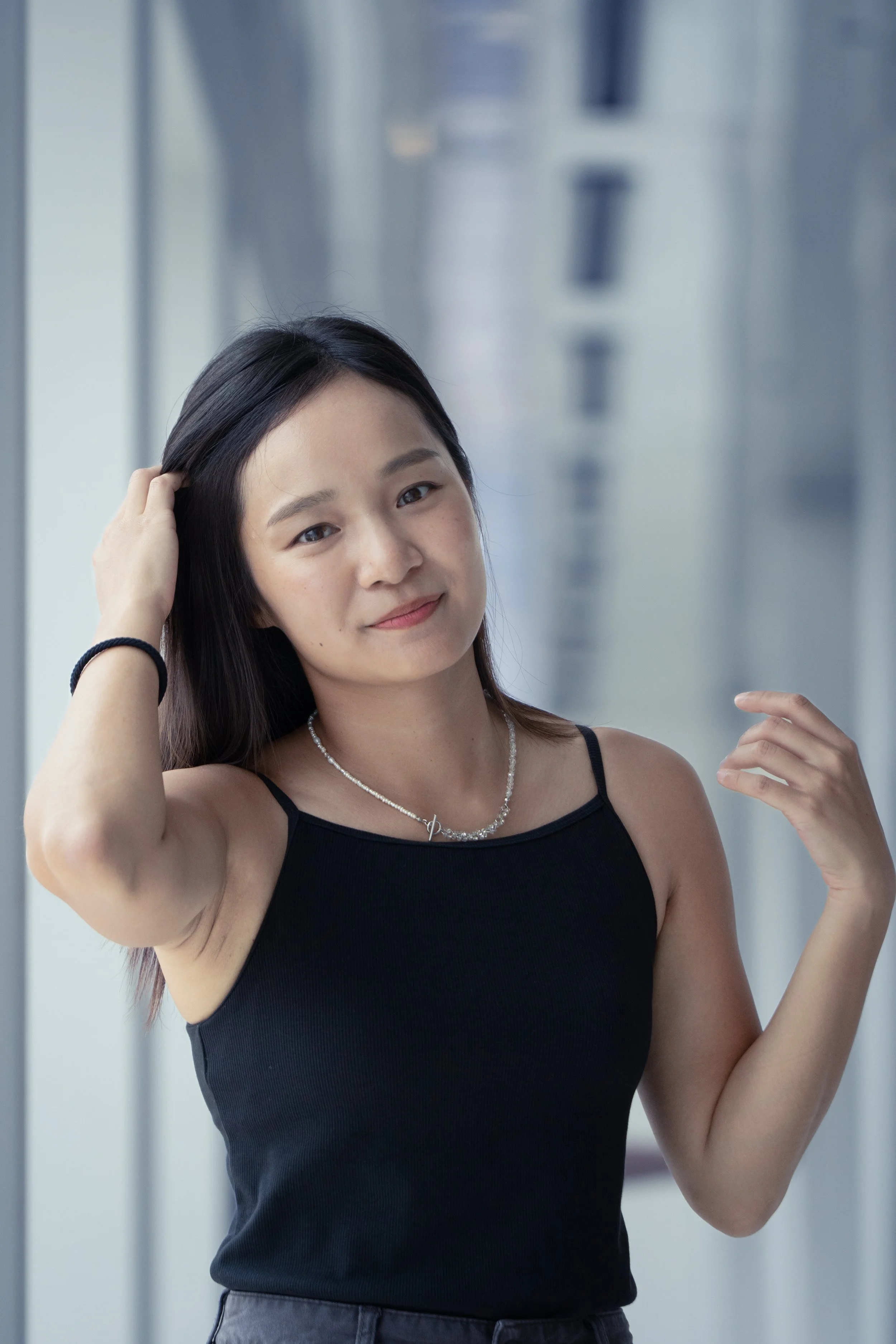 A young woman with straight dark hair wearing a black spaghetti strap top, accessorized with a silver necklace and a black bracelet, posing confidently in front of a glass window.