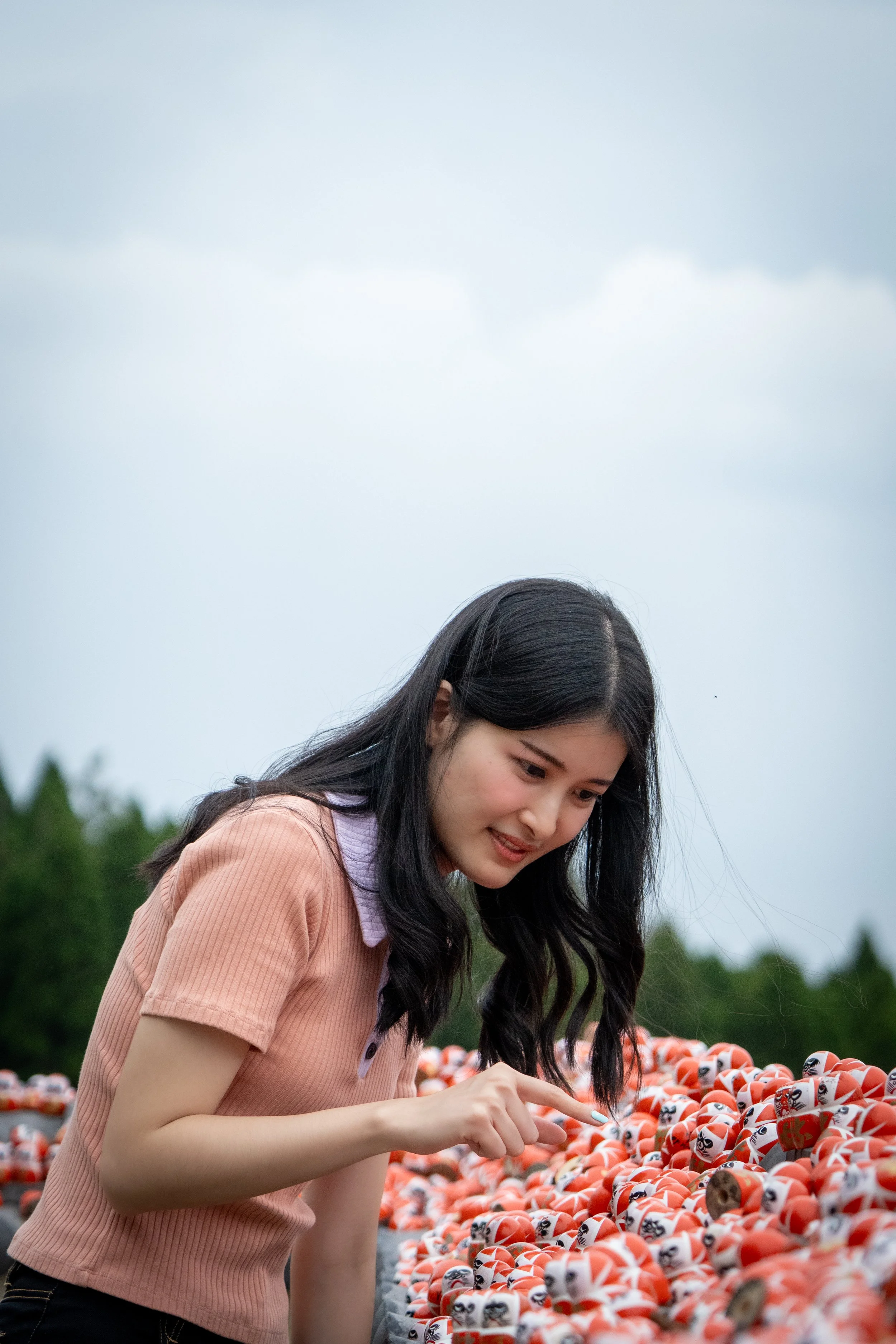 A young woman with long black hair, wearing a pink short-sleeve shirt, pointing at a row of colorful Daruma dolls outdoors on an overcast day.