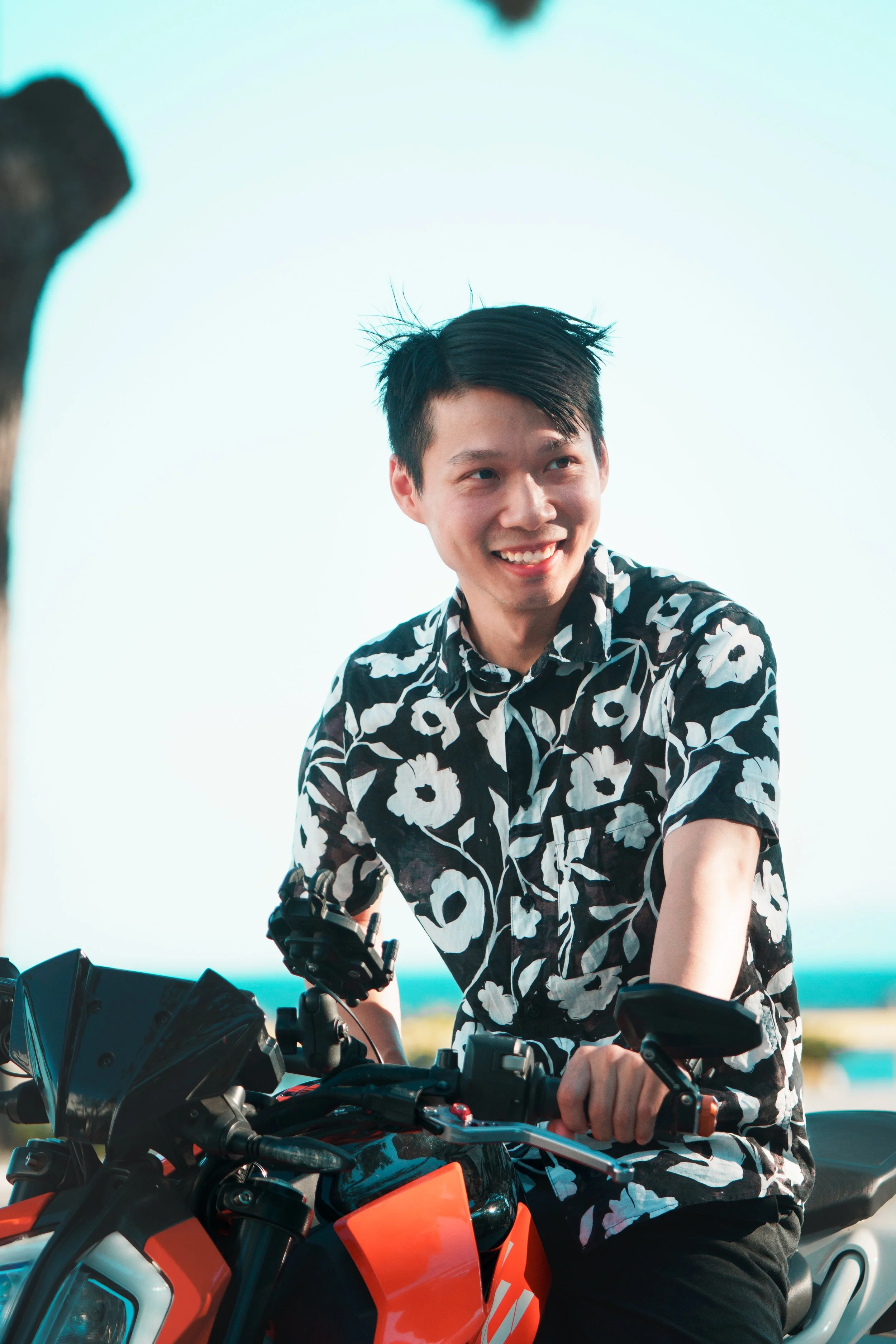A young man with short black hair smiling while sitting on a red and black motorcycle, wearing a black and white floral shirt, outdoors with a clear sky in the background.