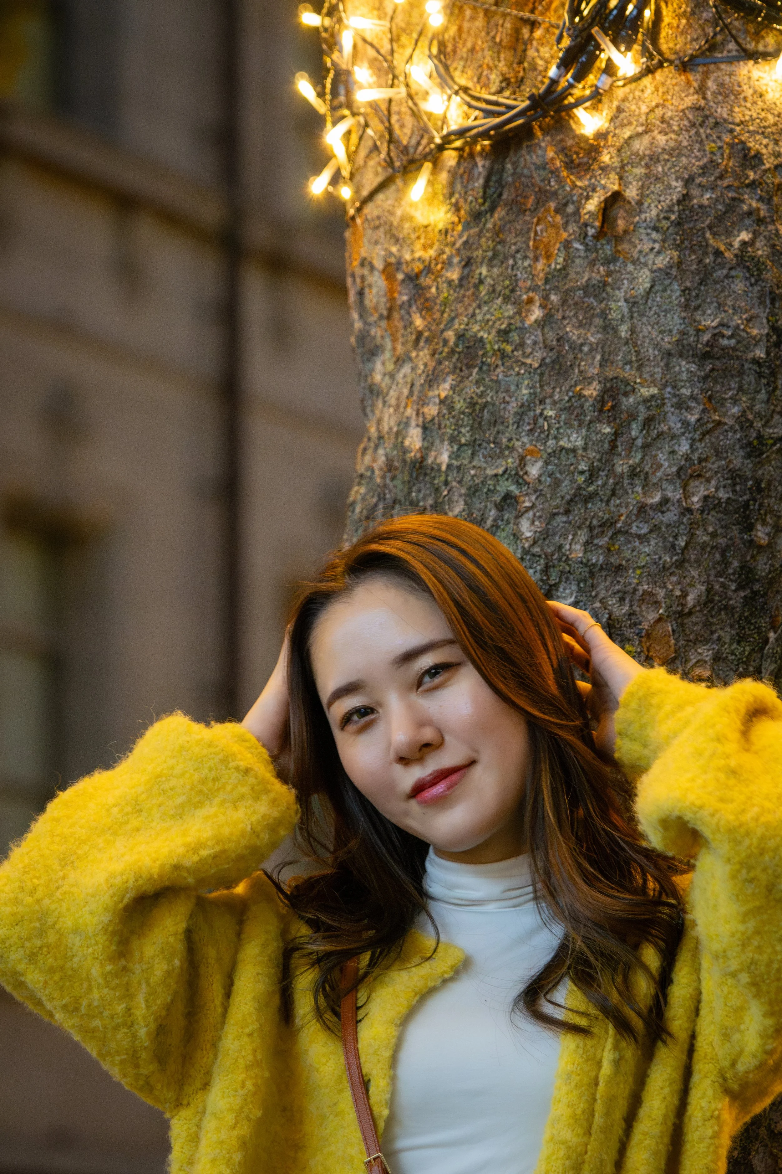 A woman in a yellow coat standing near a tree with string lights, looking at the camera.