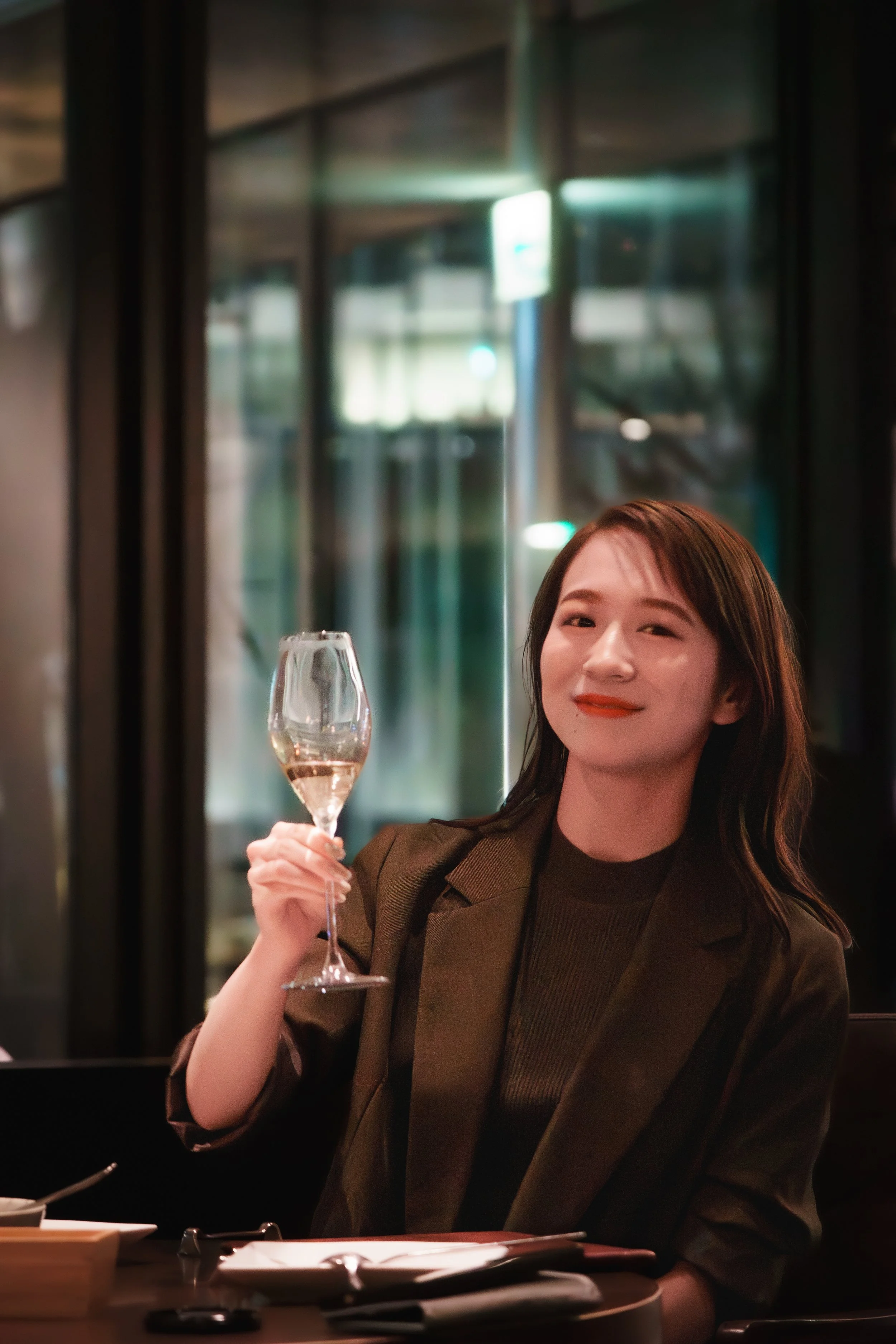 A woman with shoulder-length brown hair smiling and holding a glass of rosé wine in a restaurant or bar setting.