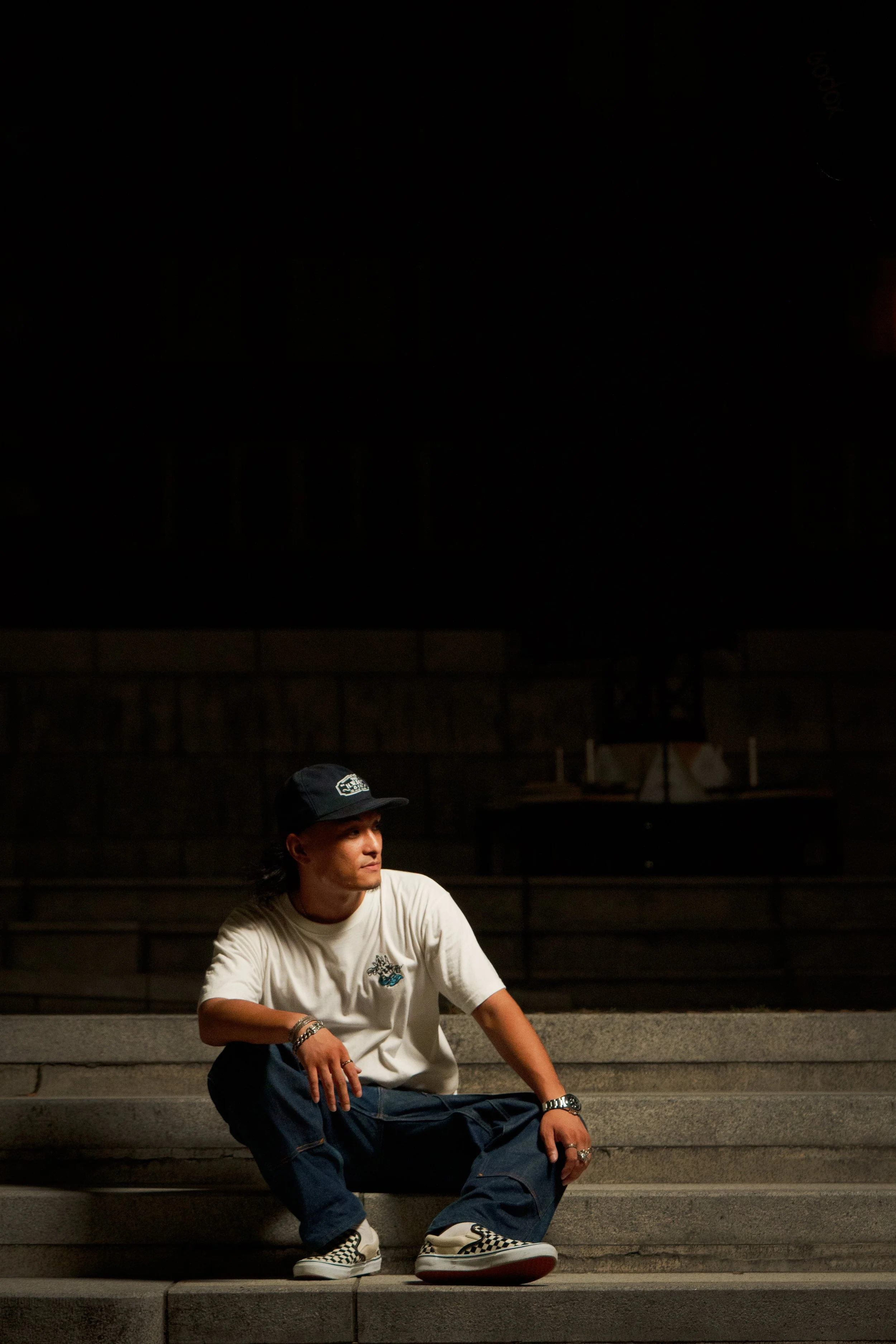 A young man sitting on steps at night, wearing a white t-shirt, dark pants, checkered shoes, a black cap, and accessories.