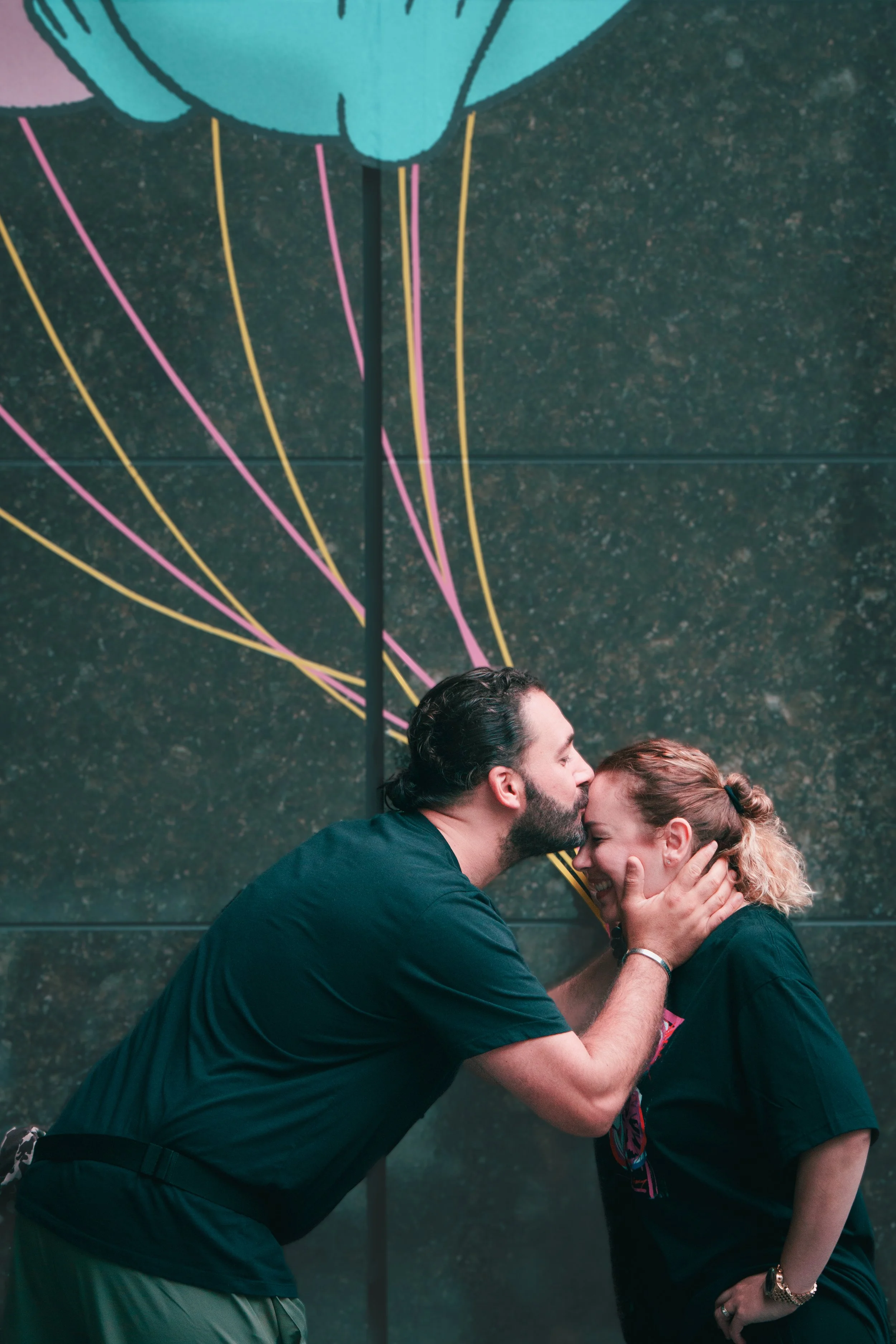 A man gives a woman a kiss on the forehead while holding her face, both smiling, against a dark wall with colorful abstract neon art.