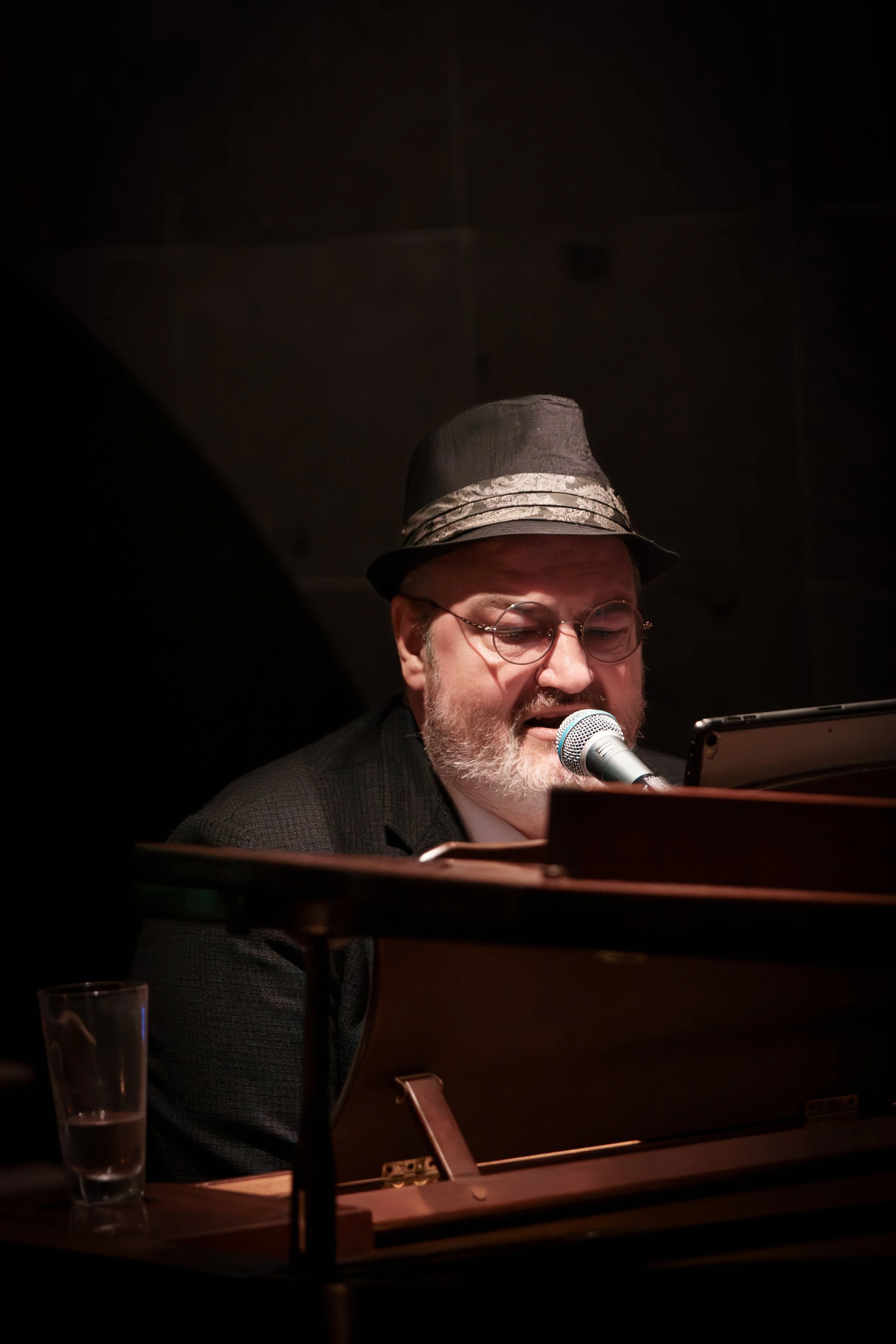 A man with glasses and a gray beard wearing a black hat and a dark jacket, singing into a microphone while playing the piano.