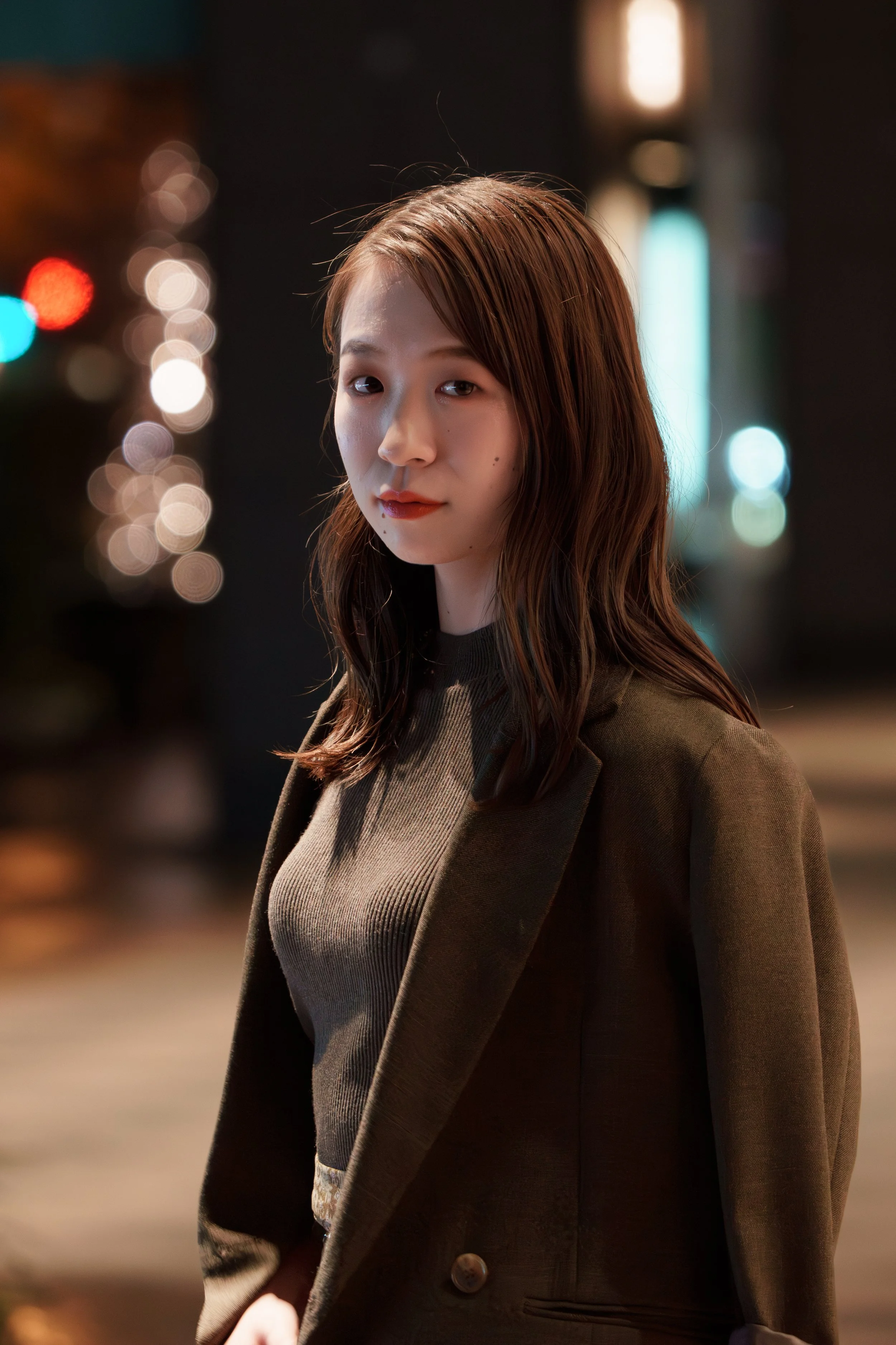 A young woman with brown, wavy hair stands outdoors at night, dressed in a dark blazer over a ribbed top, with a blurred, colorful bokeh background.