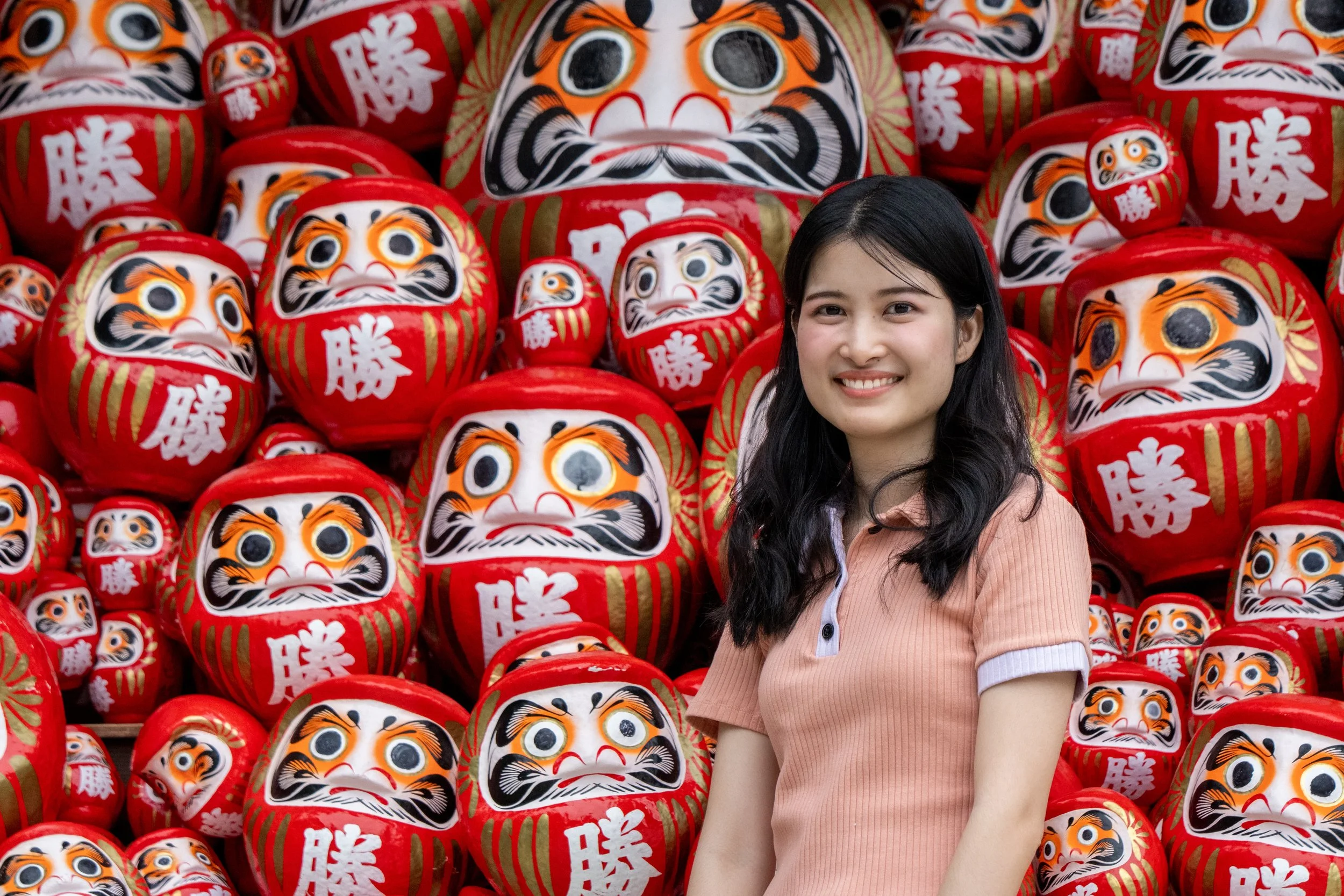 A young woman smiling in front of a collection of red Japanese Daruma dolls with white, black, and orange facial features.