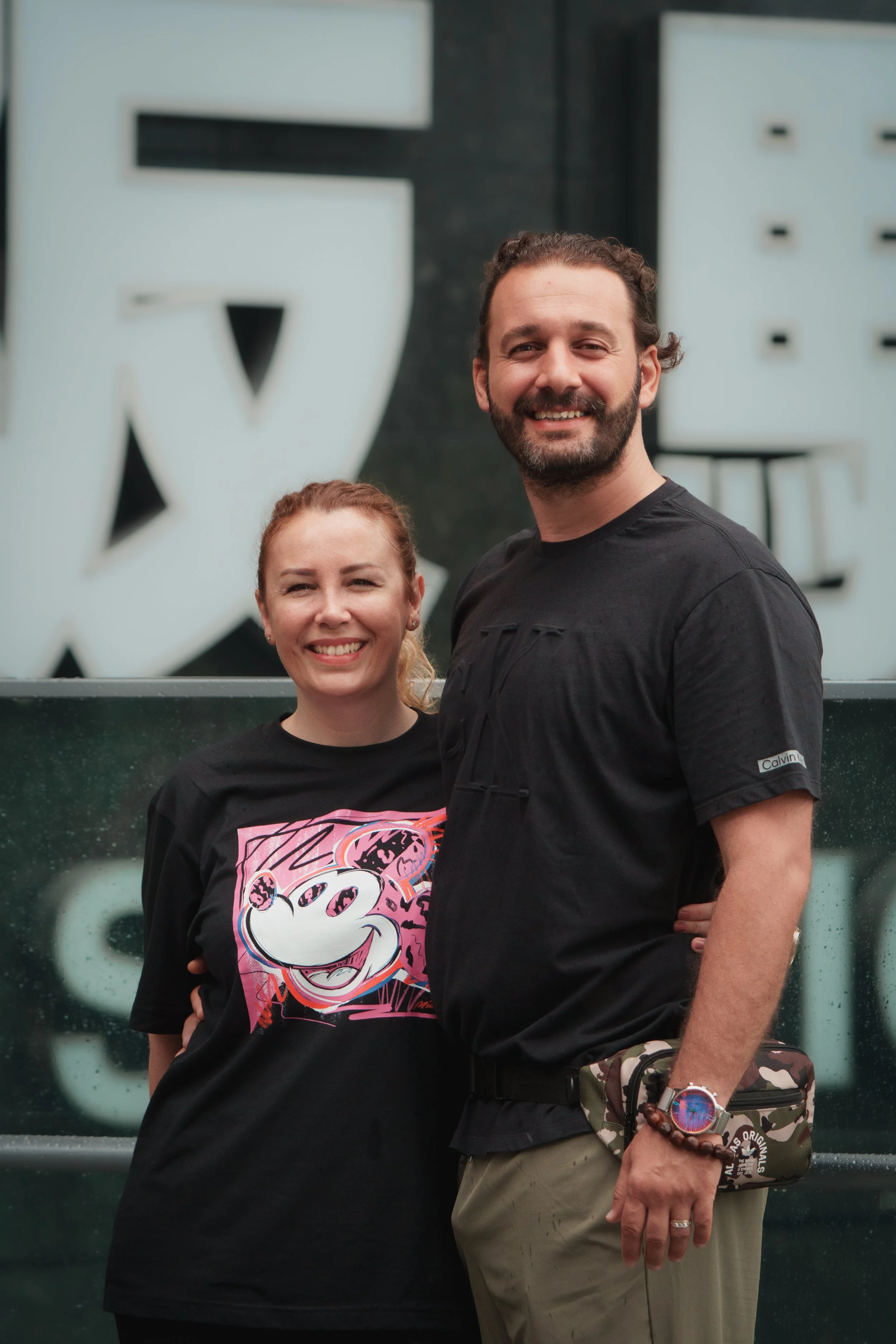 A smiling man with dark hair and beard wearing a black t-shirt and khaki pants, standing next to a smiling woman with red hair in a black t-shirt featuring a Mickey Mouse graphic, both posing outdoors in front of large white block letters.