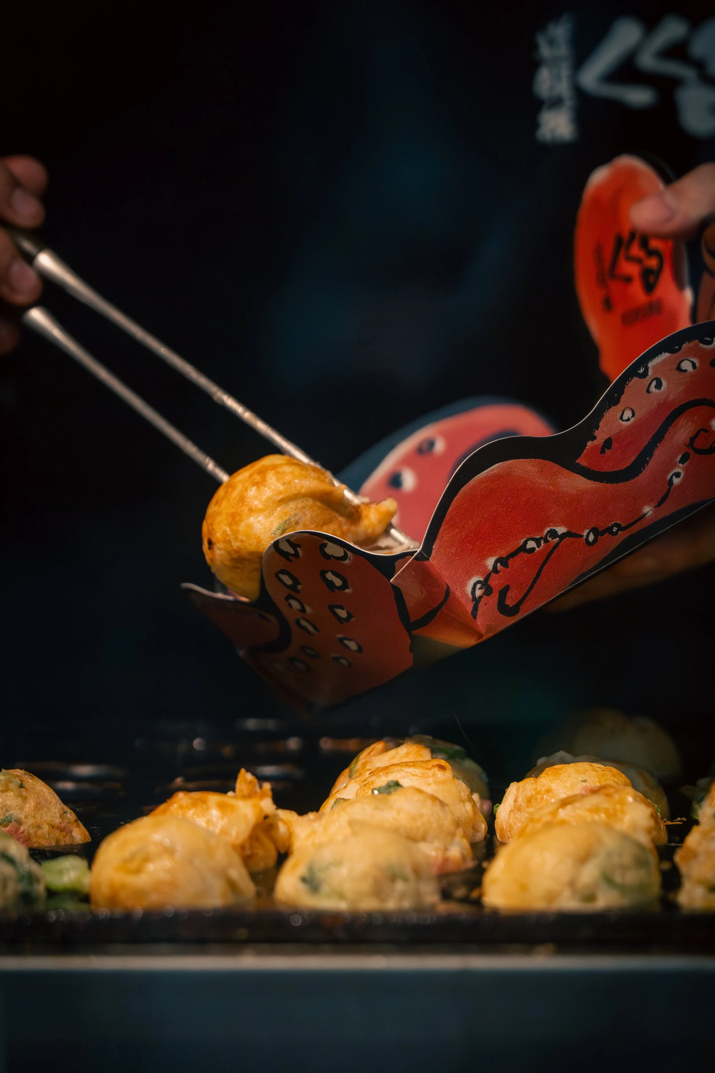 Close-up of a person using tongs to pick up a takoyaki ball from a tray, with several similar balls cooking below. The person is wearing a black apron with colorful design and text.
