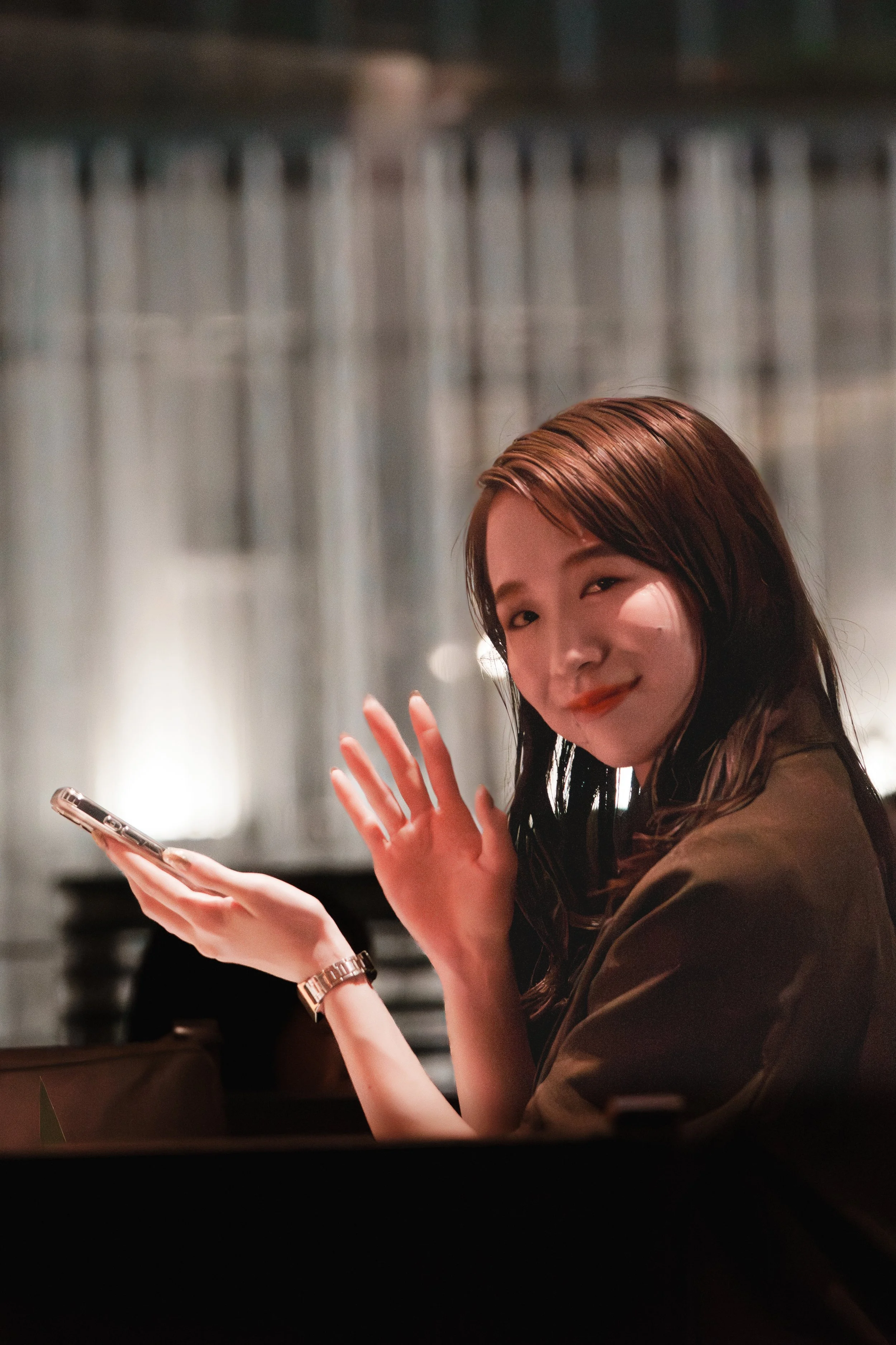 A woman with brown hair smiling and waving at the camera while holding a smartphone, sitting in a dimly lit indoor setting.