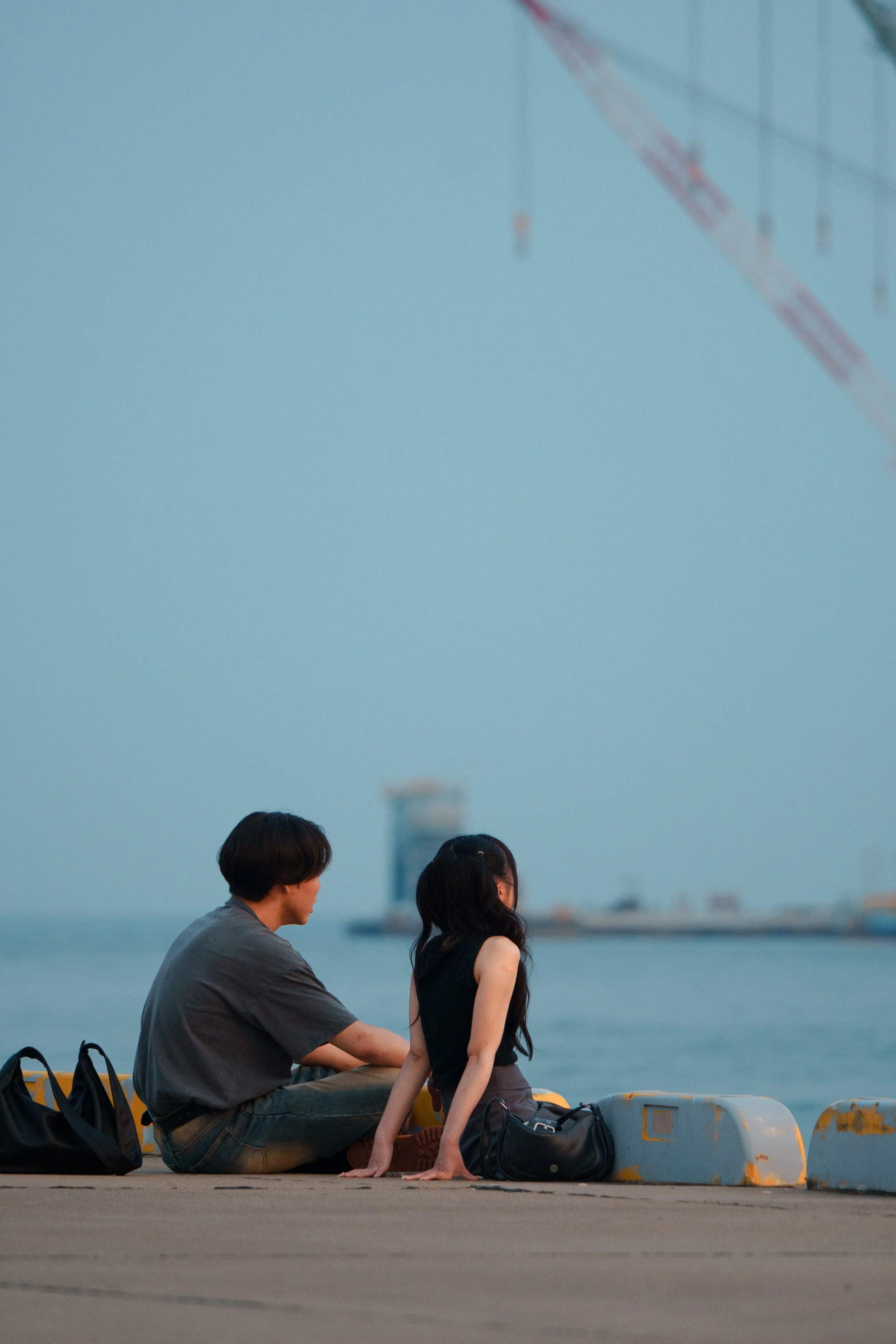 A young man and woman sitting on a pier by the water, facing each other during sunset, with bags placed beside them and a blurred city skyline and crane in the background.
