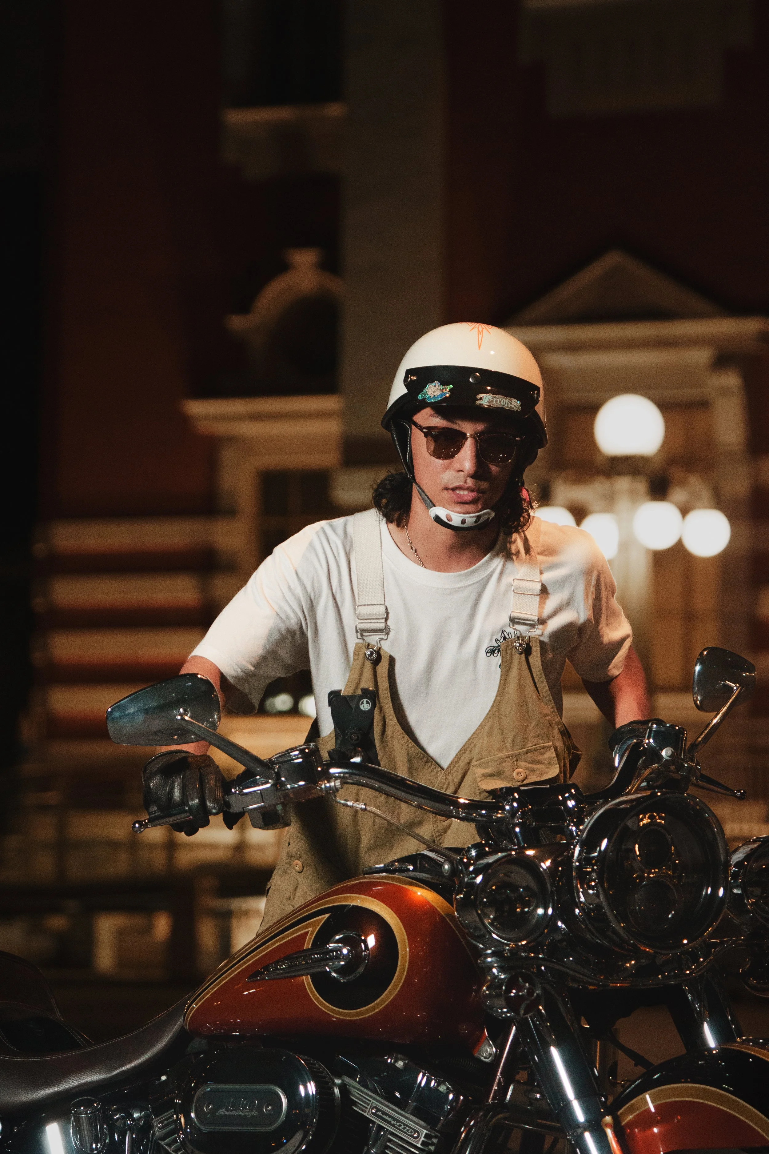 A young man wearing sunglasses, a white helmet, a white t-shirt, and tan overalls riding a black motorcycle at night in an urban setting.