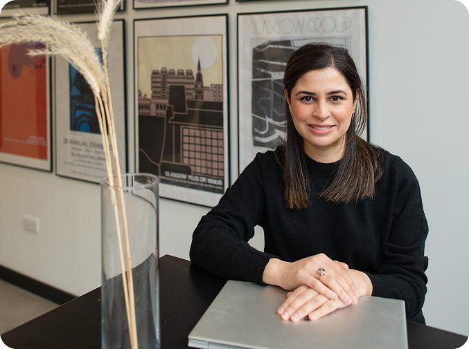 A young woman with dark hair and a black sweater sitting at a table in front of a laptop, smiling. Behind her are framed posters on a white wall.