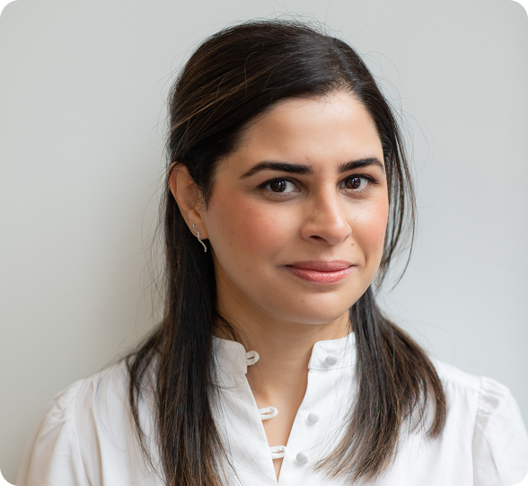 A woman with dark brown hair, wearing a white blouse and small earrings, smiling slightly, standing against a neutral background.