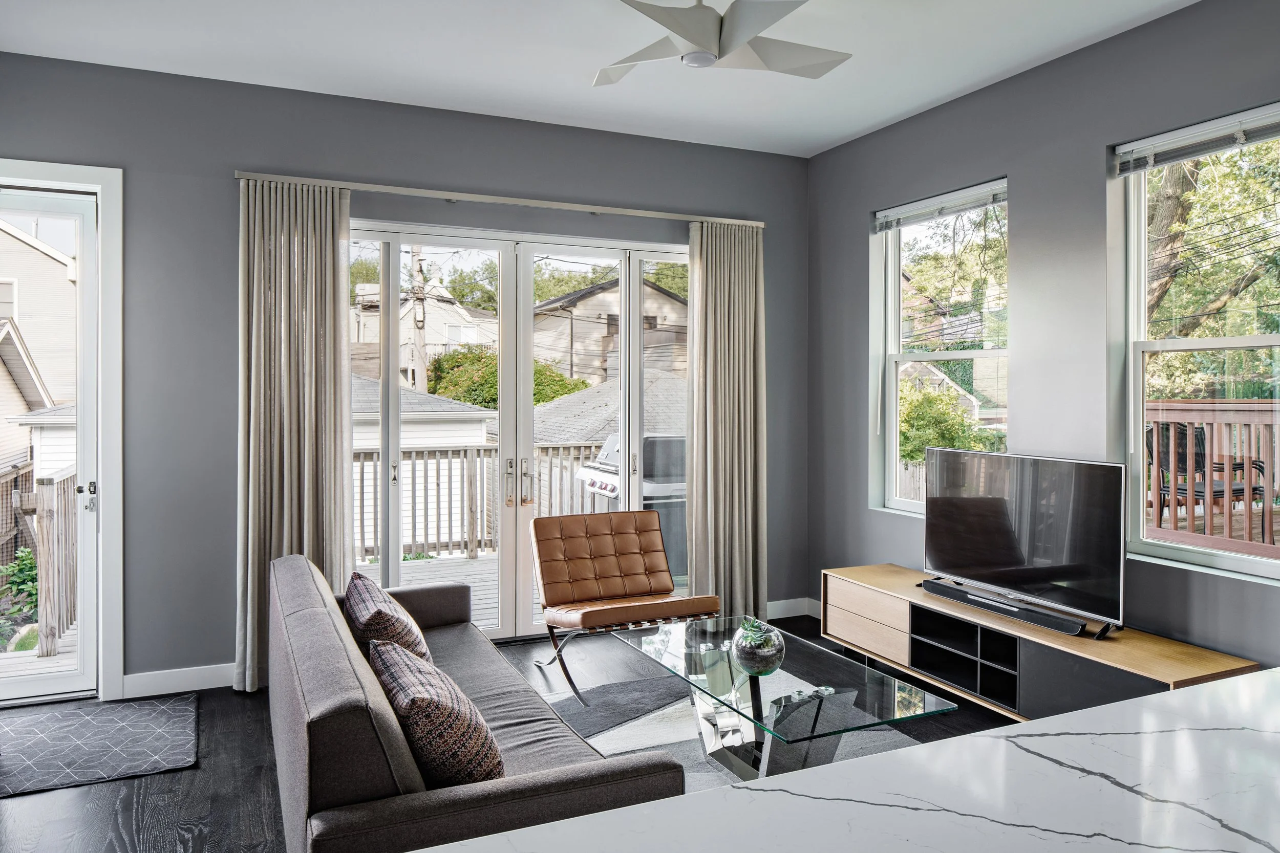 Living room with large windows, a gray sofa with pillows, a glass coffee table, a wooden TV stand with a flat-screen TV, and a brown leather chair. Outside view shows houses, trees, and a deck with a barbecue grill.