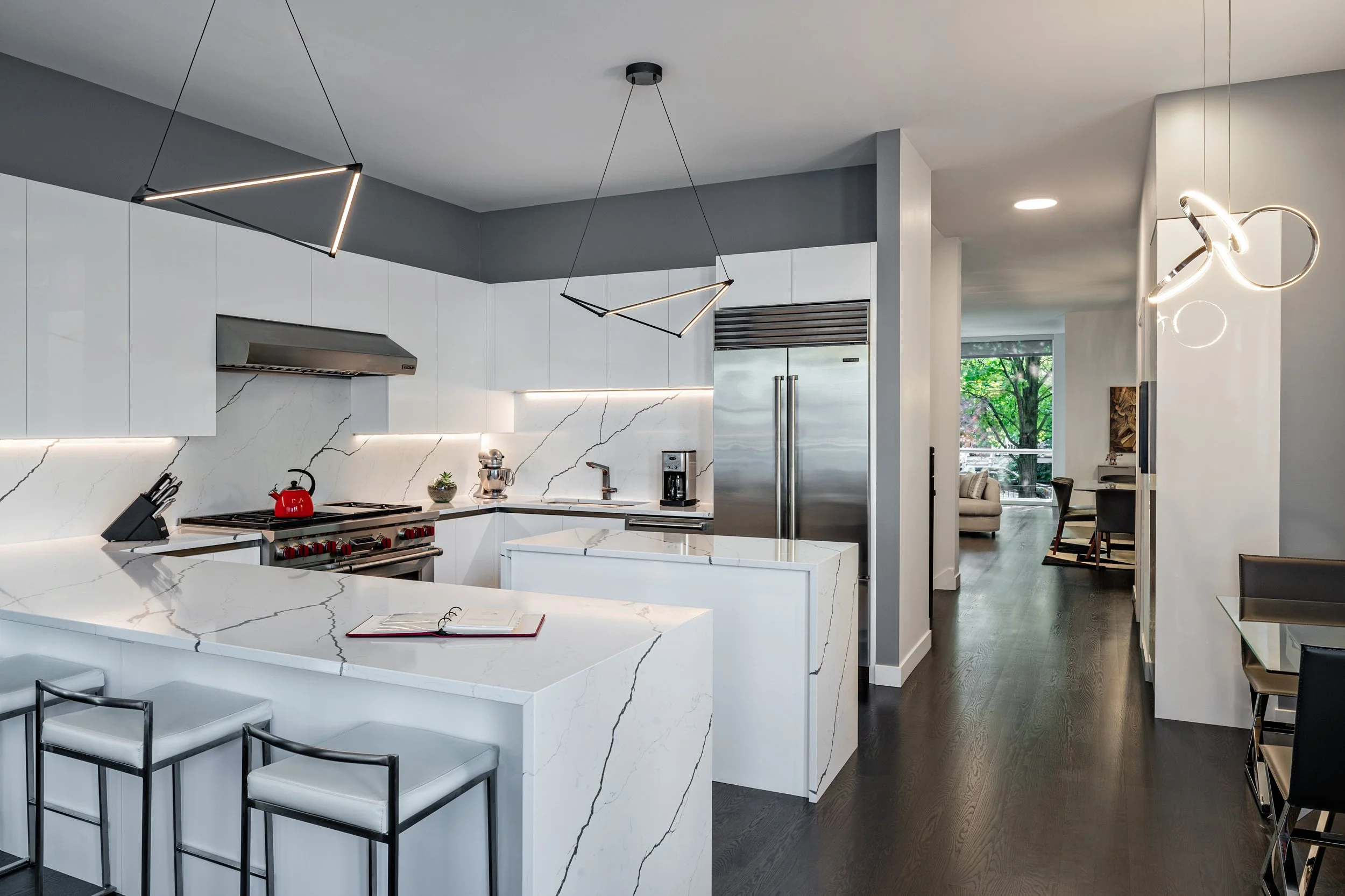 Modern kitchen with white marble countertops, white cabinets, stainless steel appliances, black pendant lights, and dark hardwood flooring. A glimpse into an adjacent living area with large windows and outdoor greenery is visible.