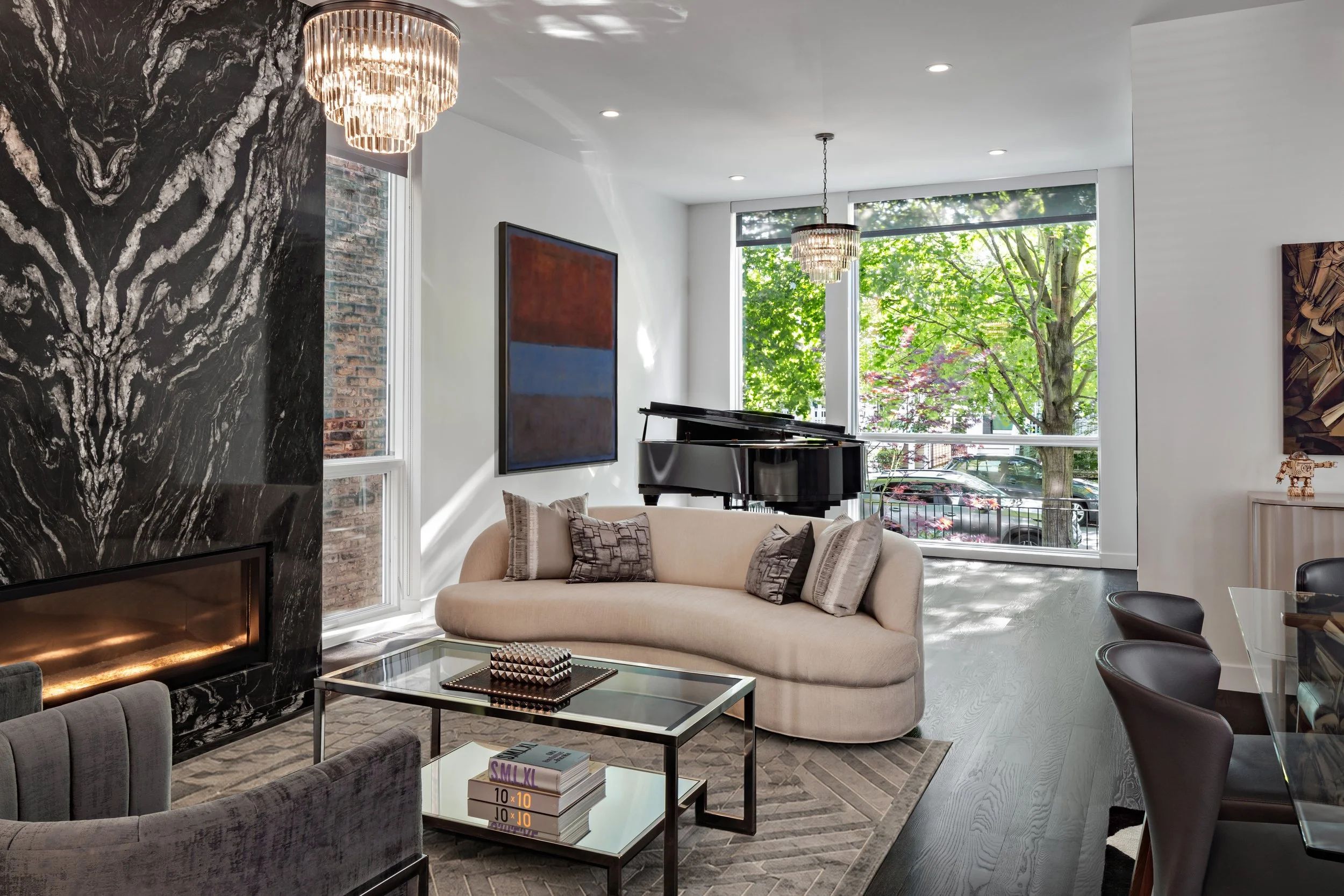 A modern living room with a black marble fireplace, a beige sofa with decorative pillows, a glass coffee table with books, a black grand piano near large windows, and leafy trees visible outside. There are two chandeliers hanging from the ceiling wit