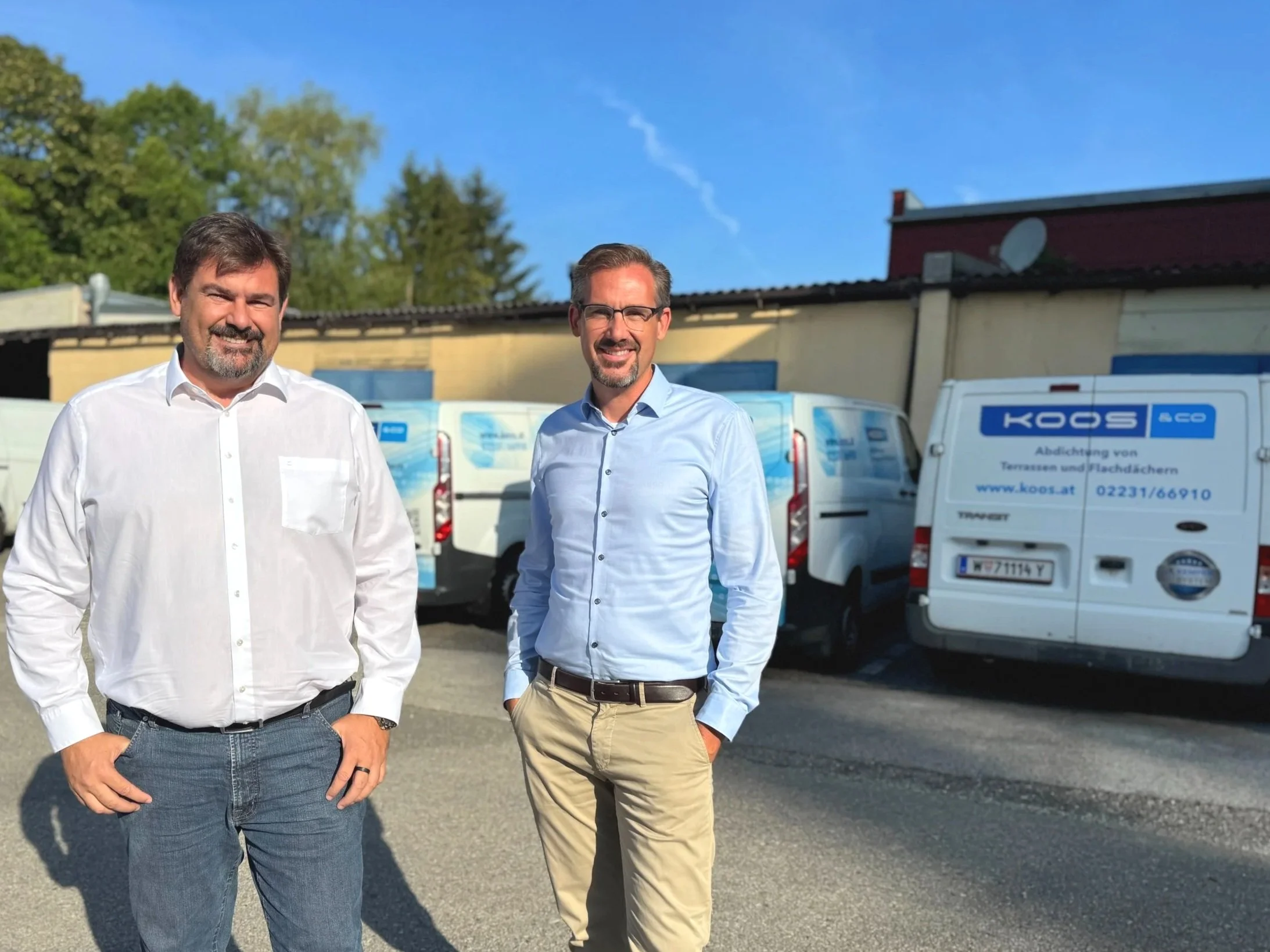 Stefan Steinbichler und Andreas Steinbichler, Geschäftsführer der Firma Koos, stehen im Freien vor Firmenfahrzeugen. Hintergrund mit Bäumen und Gebäude, blauer Himmel. Beide Männer tragen Hemden, lächeln und schauen in die Kamera.