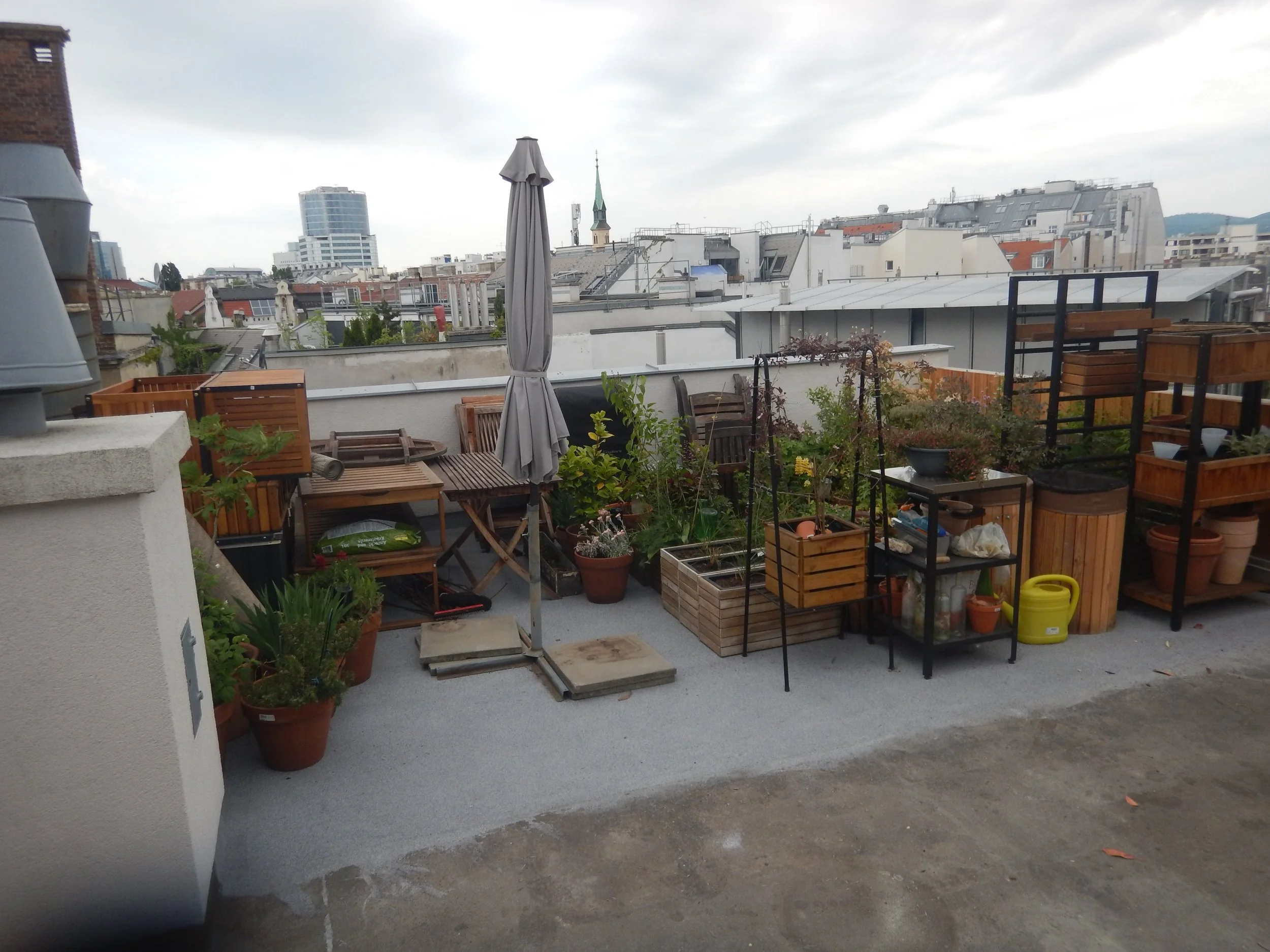 Ansicht einer Terrasse mit vielen Möbelstücken, Pflanzgefässen, Einrichtungsgegenständen und Zugeklapptem Schirm.