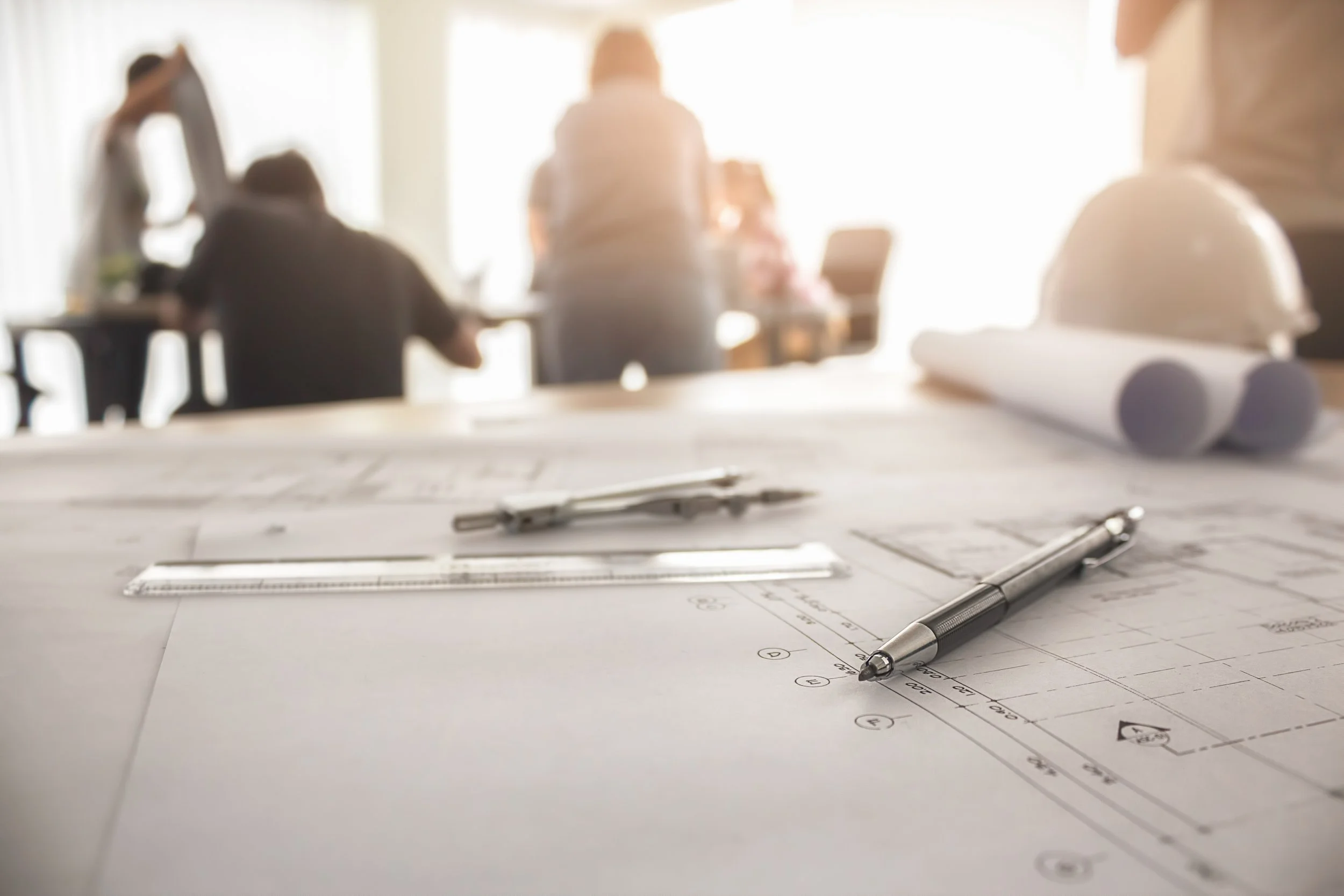 Dessins d'architecture, une règle, un stylo et un casque de sécurité posés sur des plans sur une table de travail, avec des ouvriers et un ingénieur en arrière-plan dans un bureau lumineux.