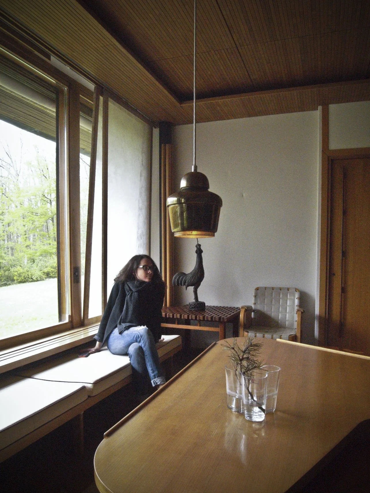 Une femme assise sur un long coussin près d'une grande fenêtre, dans une pièce avec un plafond en bois, une table en bois, une lampe suspendue en laiton avec une sculpture de coq en dessous, et deux verres avec une plante dans un vase transparent.