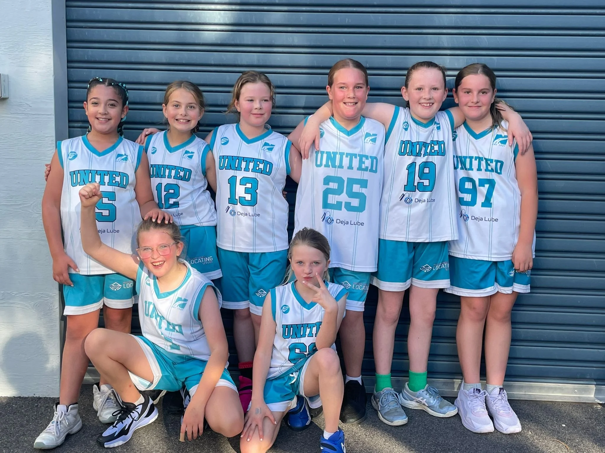 A group of young girls in matching blue and white basketball uniforms posing in front of a metal building shutter. They are smiling and some have their arms around each other.