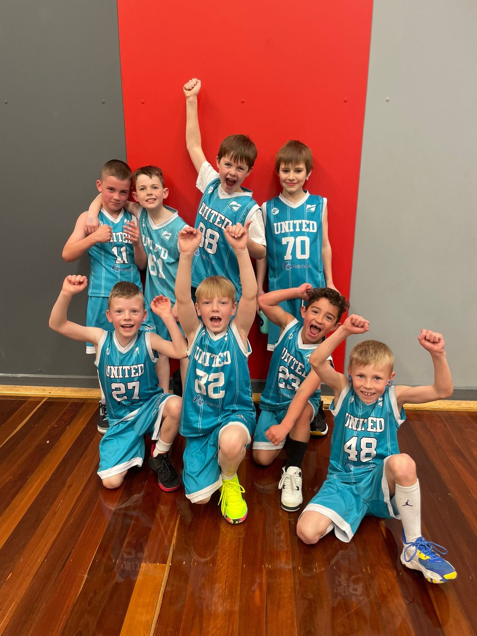 A group of young boys in blue basketball uniforms posing for a team photo indoors with a red wall behind them. They are smiling and making victory poses.