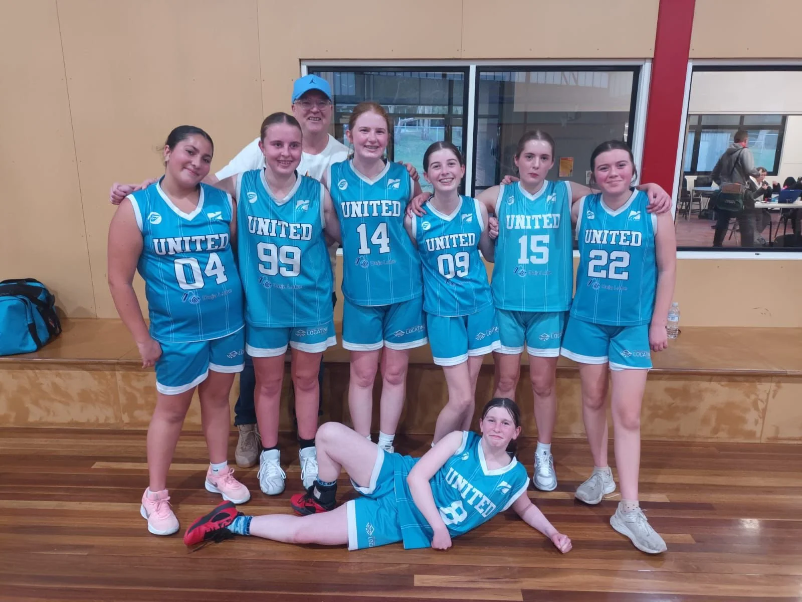 A group of young female basketball players and one adult man posing inside a gym. The players are wearing matching blue jerseys with 'UNITED' written on them, and each has a different number. They are standing and sitting in front of a window, with a man in a white shirt, black pants, and a blue cap standing behind them. One girl is lying on the floor in front of the group.