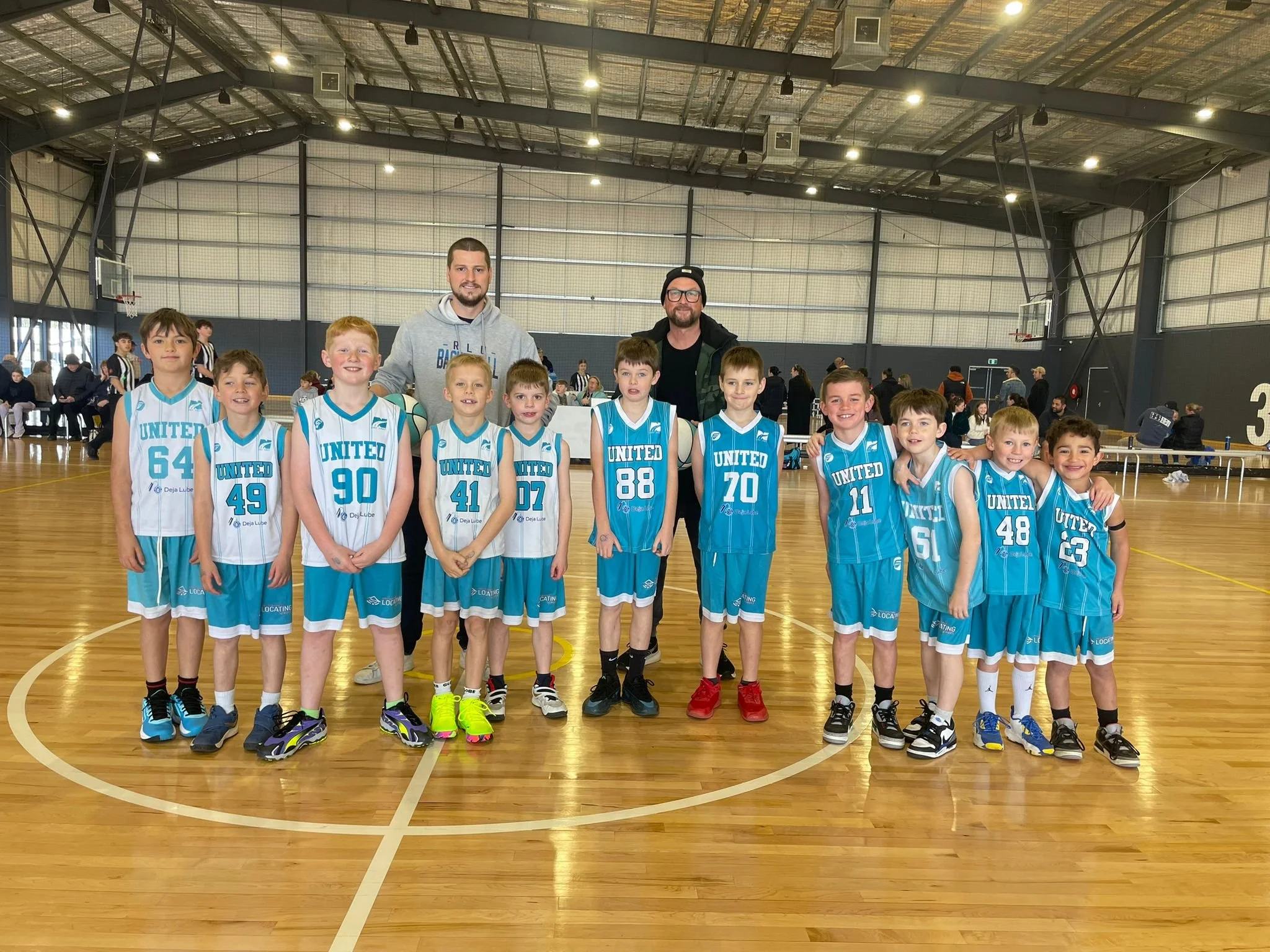 A group of young boys in blue and white basketball uniforms standing together with two adult men inside a large indoor gymnasium. The boys are smiling and appear to be part of a basketball team. The gym has a high ceiling with lighting and sports equipment, including basketball hoops.