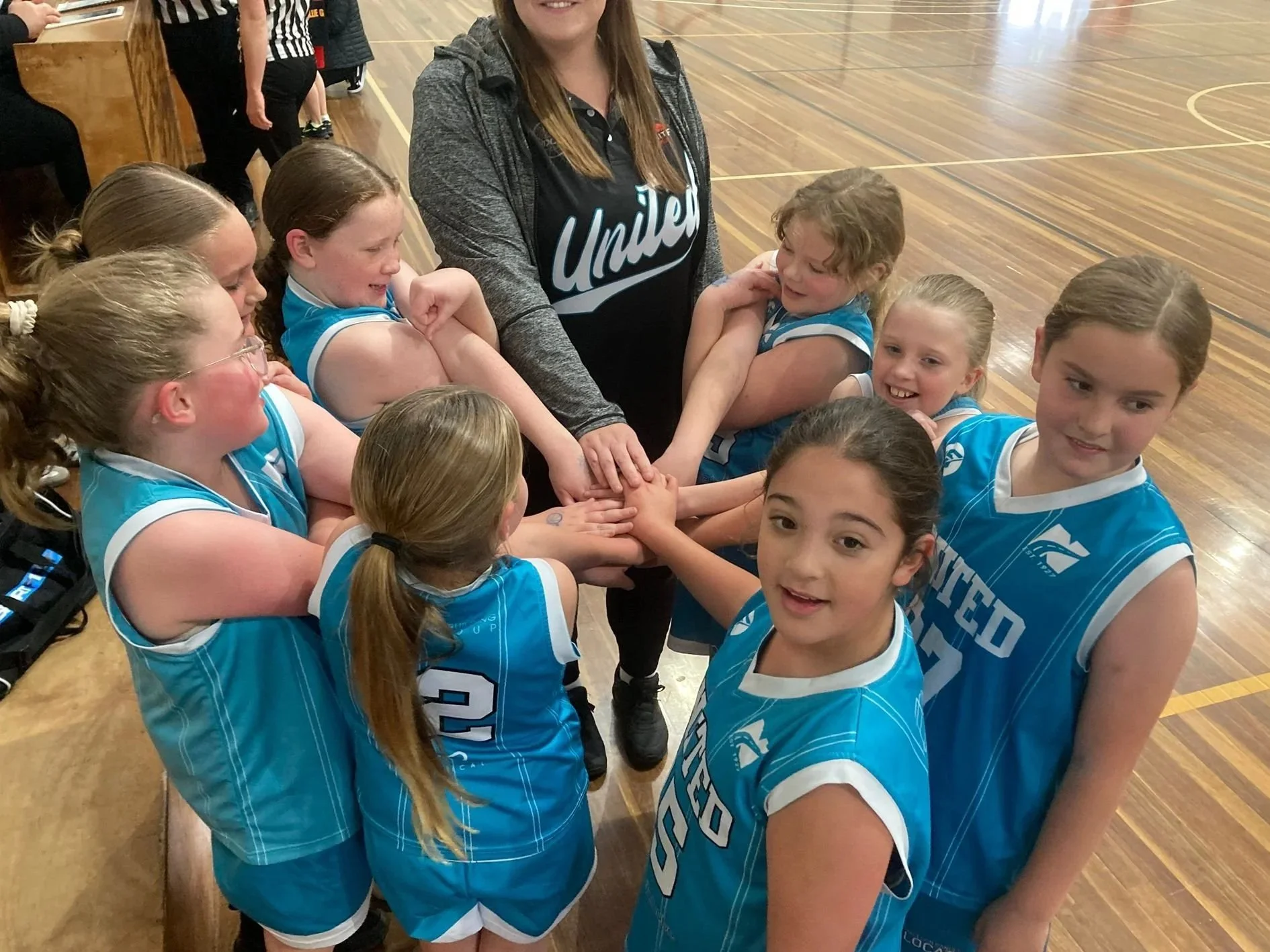 A group of young girls in blue sports uniforms standing in a circle with an adult woman in a black shirt and gray jacket in the center, inside a gymnasium.